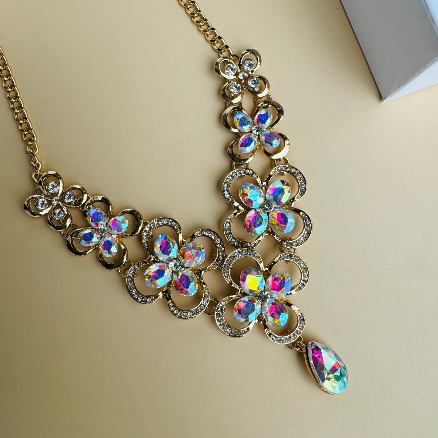 Gold statement necklace with iridescent floral-shaped gemstones and a teardrop pendant, displayed on a white mannequin bust. The intricate design sparkles with multicolored reflections, creating a luxurious and elegant look