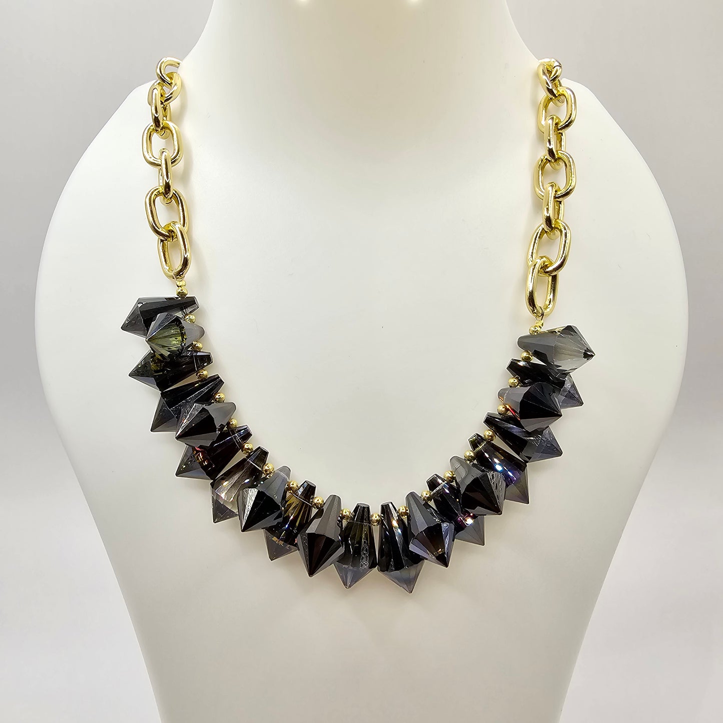 A bold and edgy statement necklace featuring faceted black crystal spikes with gold-tone accents, strung on a chunky gold chain, displayed on a mannequin bust.