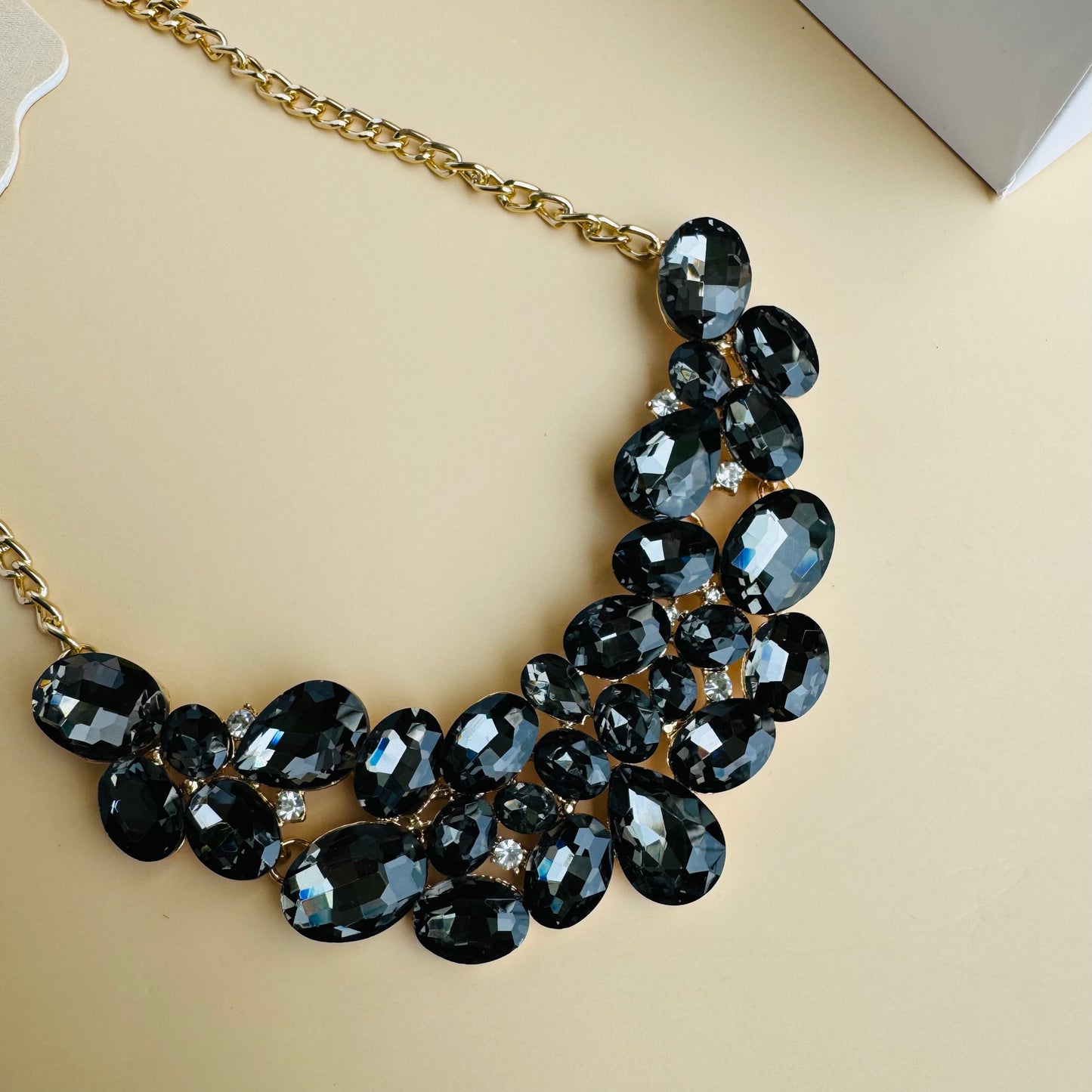 Elegant statement necklace with black and smoky crystal stones on a gold chain, displayed on a white mannequin bust. Branded Lush 24 packaging in the background adds a premium touch.