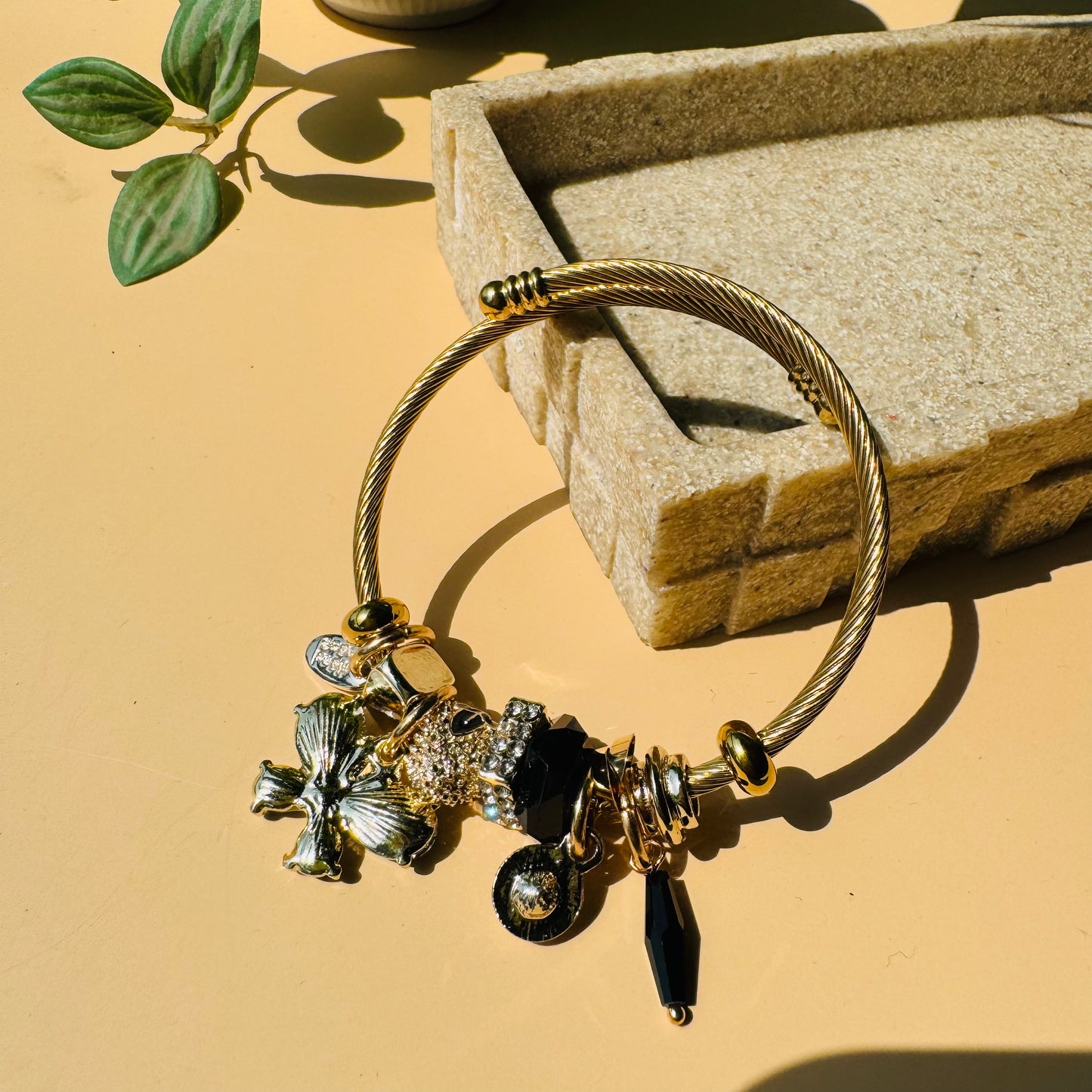 A Pandora-style charm bracelet with a gold-toned wire bangle and a collection of charms in mixed metals, including a silver-toned flower, a pave crystal bead, and black and gold accents. Likely available in Mumbai, Maharashtra, India.