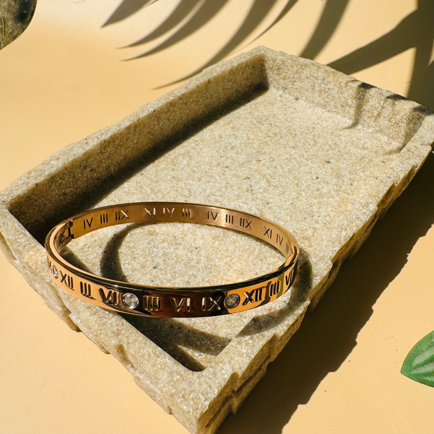 Authentic gold roman numerals Bracelet. The bracelet is showcased on a textured stone-like block with soft lighting and a glimpse of plant life.