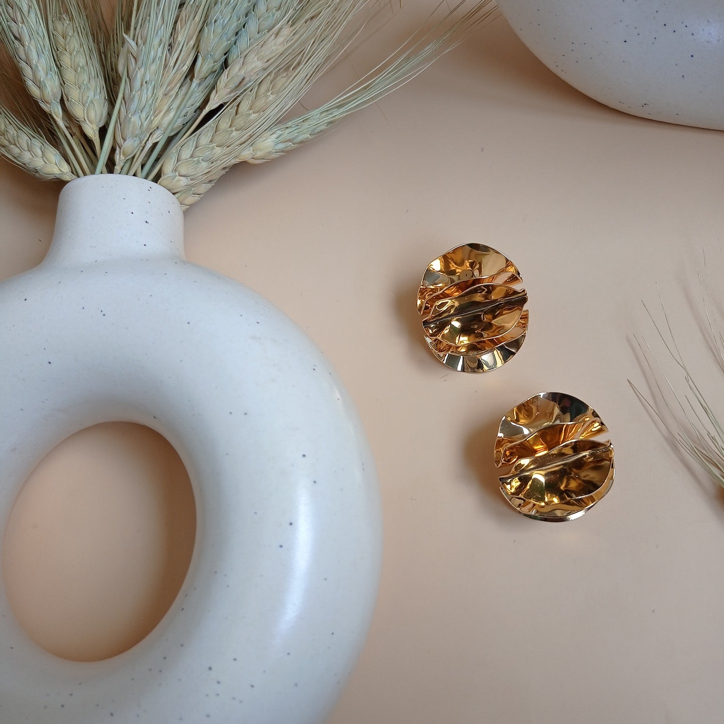 Elegant gold textured stud earrings with a sculptural, crinkled design, displayed on a neutral beige background with ceramic and dried floral elements