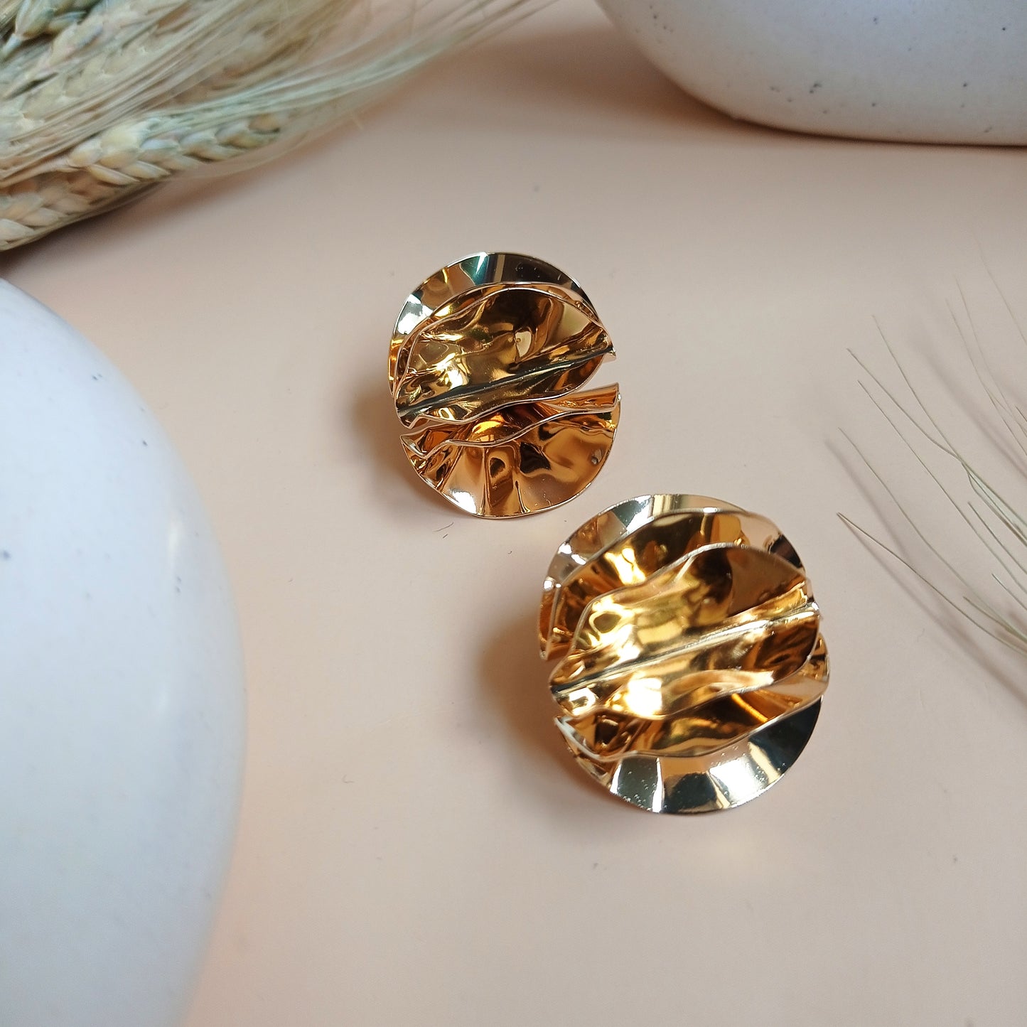 Elegant gold textured stud earrings with a sculptural, crinkled design, displayed on a neutral beige background with ceramic and dried floral elements