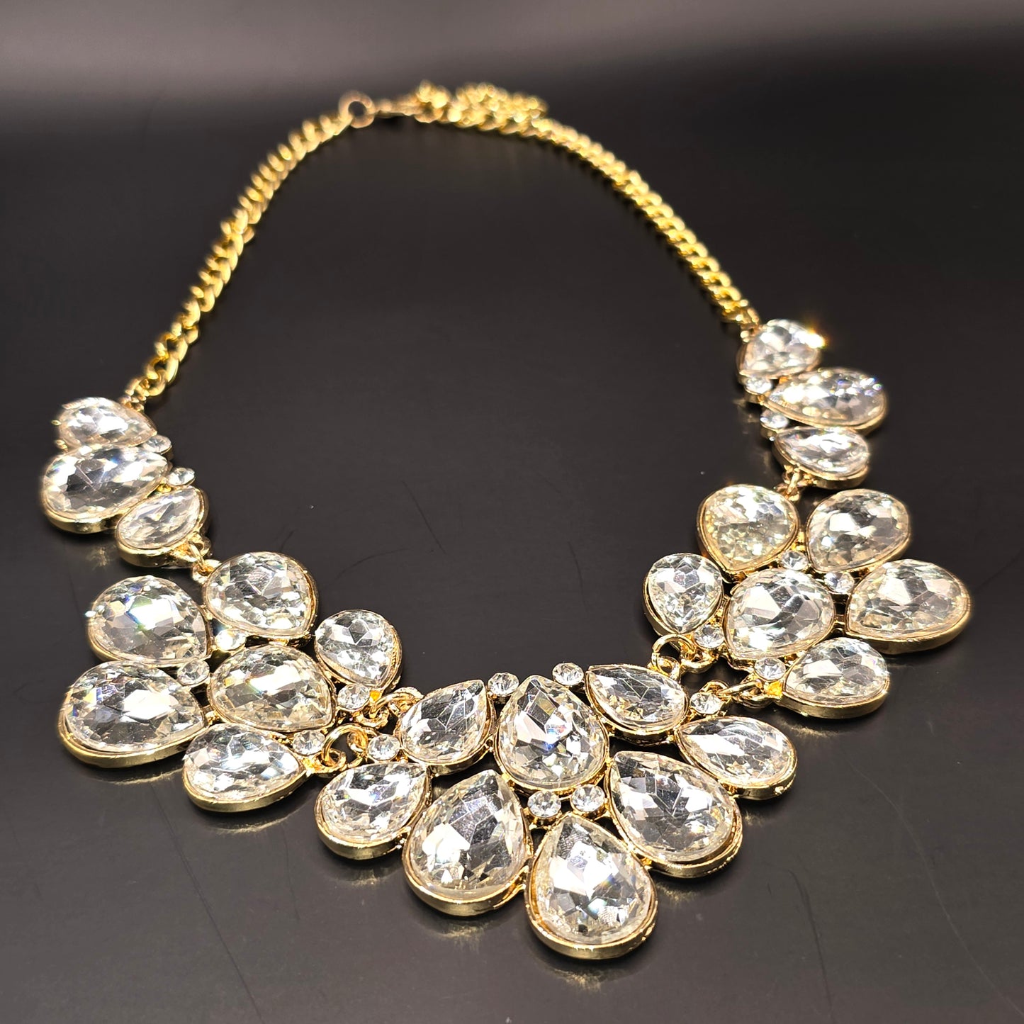 A stunning gold-tone statement necklace featuring an array of clear, faceted teardrop-shaped crystals arranged in a floral-inspired design, elegantly displayed on black satin fabric inside a jewelry box.