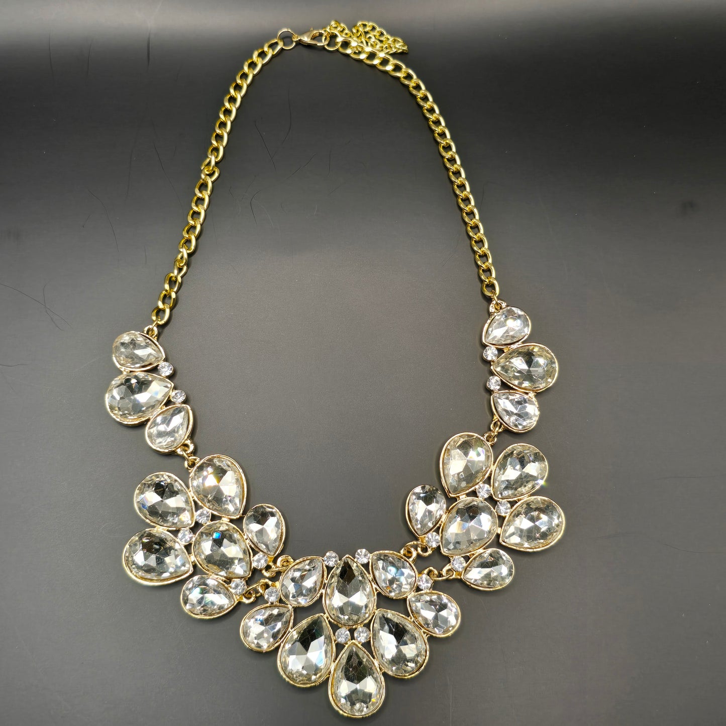 A stunning gold-tone statement necklace featuring an array of clear, faceted teardrop-shaped crystals arranged in a floral-inspired design, elegantly displayed on black satin fabric inside a jewelry box.