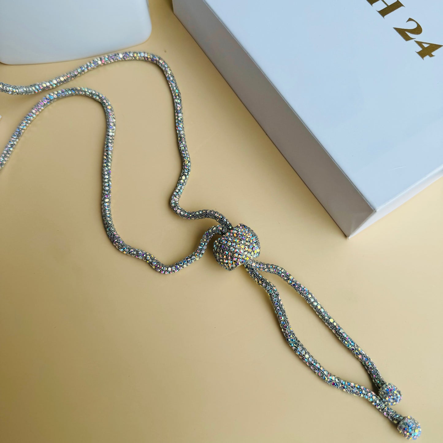 Alt text: A sparkling crystal lariat necklace displayed on a white bust stand. The necklace features a long, shimmering chain with a central crystal ball that secures two dangling strands, each ending in a smaller crystal-encrusted bead. The background includes Lush 24 branded packaging and lush green plants.