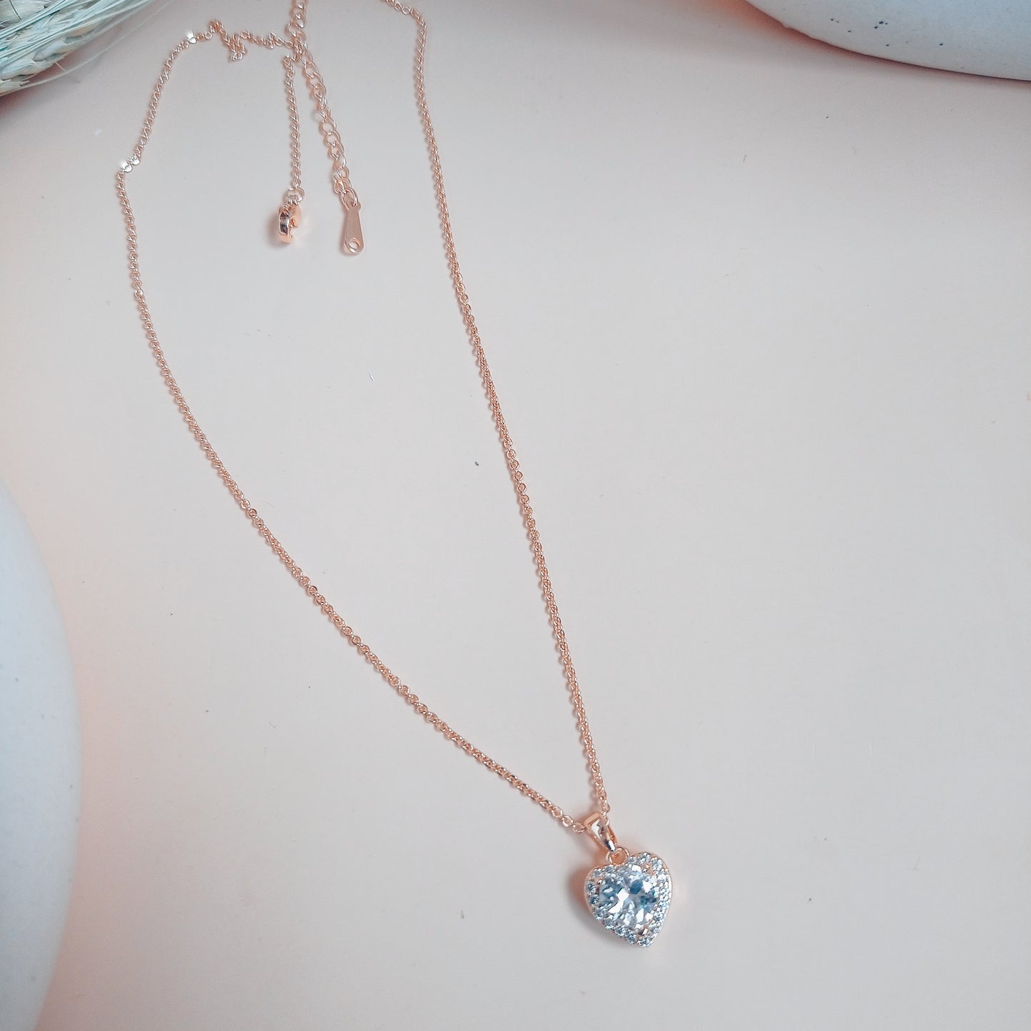 Rose gold necklace with a heart-shaped blue gemstone pendant, surrounded by sparkling white stones, set against a neutral background