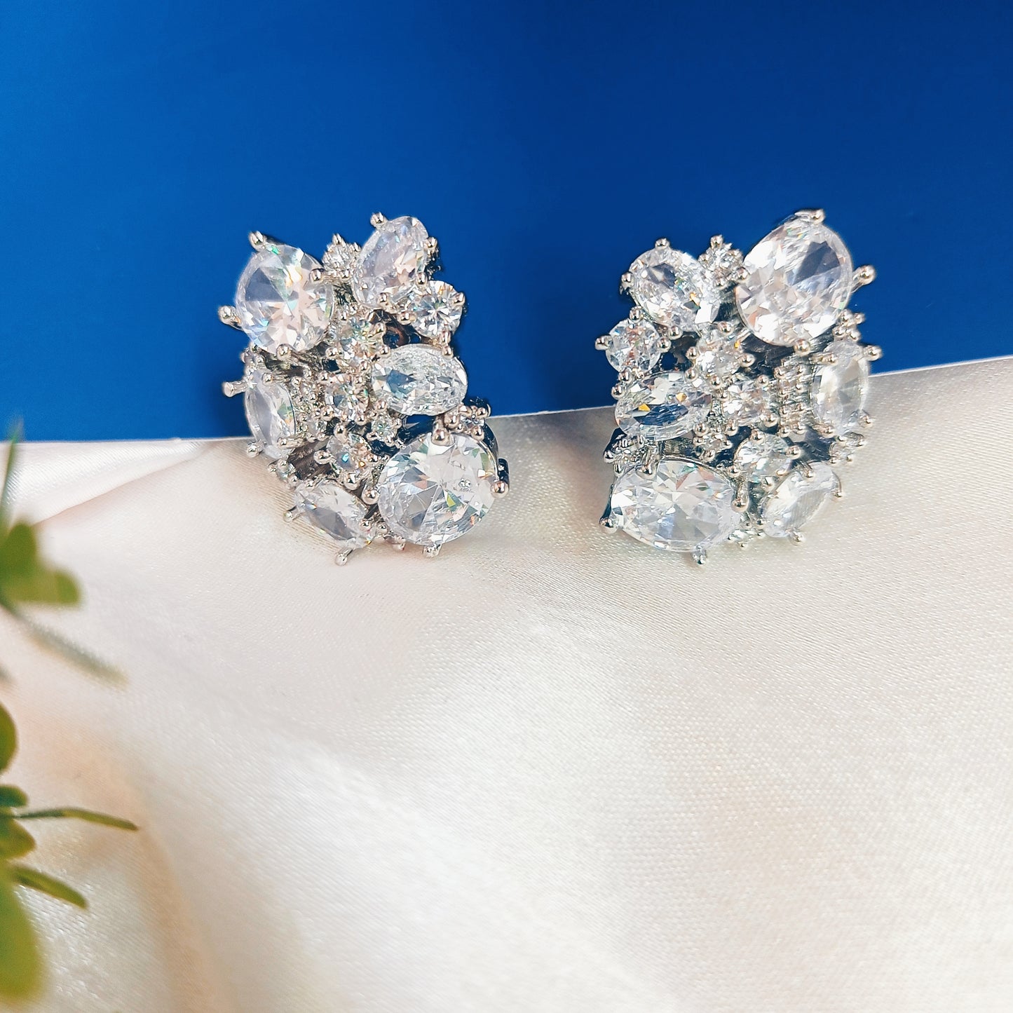 Elegant silver-tone floral cluster earrings adorned with sparkling cubic zirconia stones, arranged in a delicate, intricate design. Set against a soft, white satin background, these statement earrings exude luxury and sophistication, perfect for special occasions.