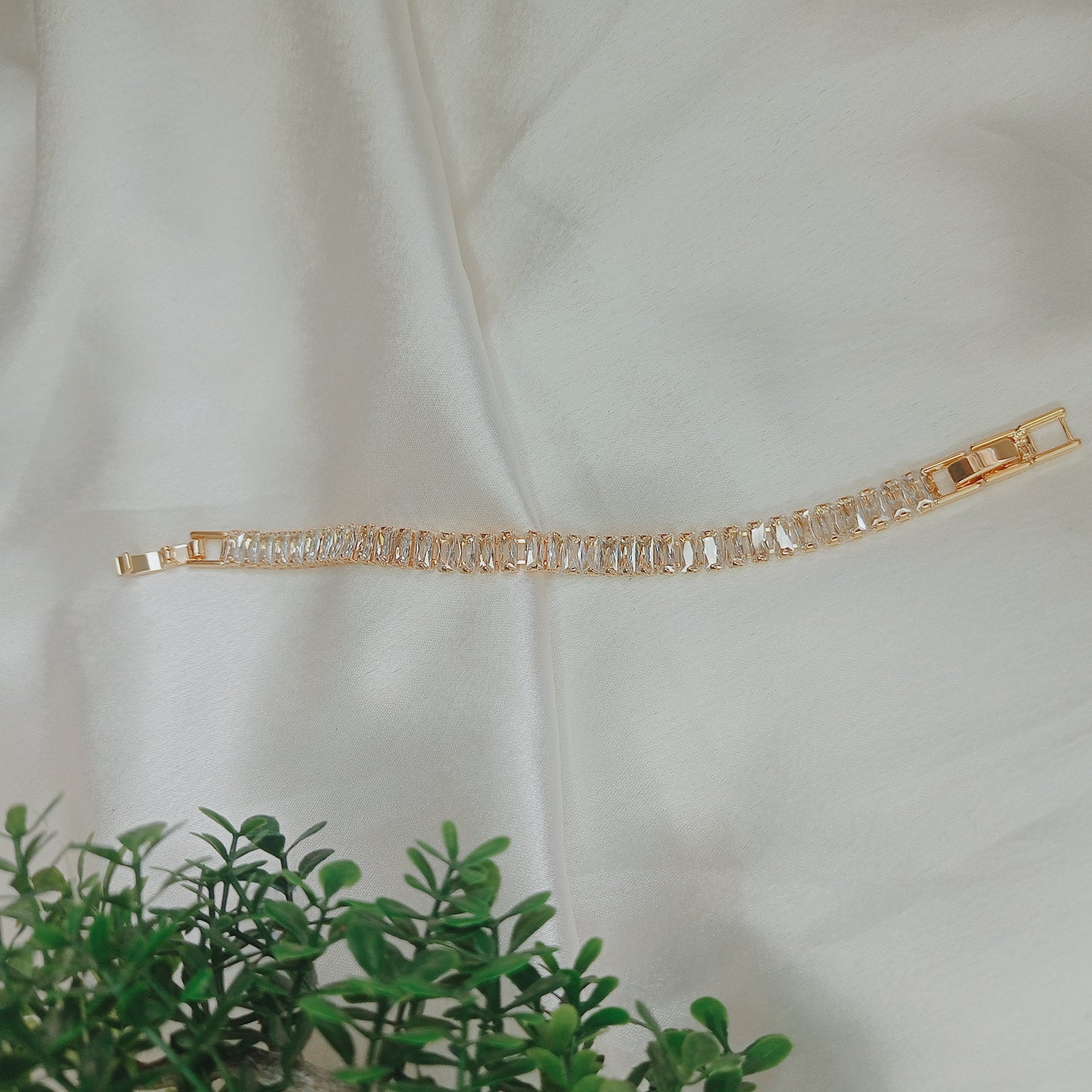 Elegant gold bracelet with baguette-cut clear gemstones displayed on a soft white satin fabric, with a touch of greenery in the foreground