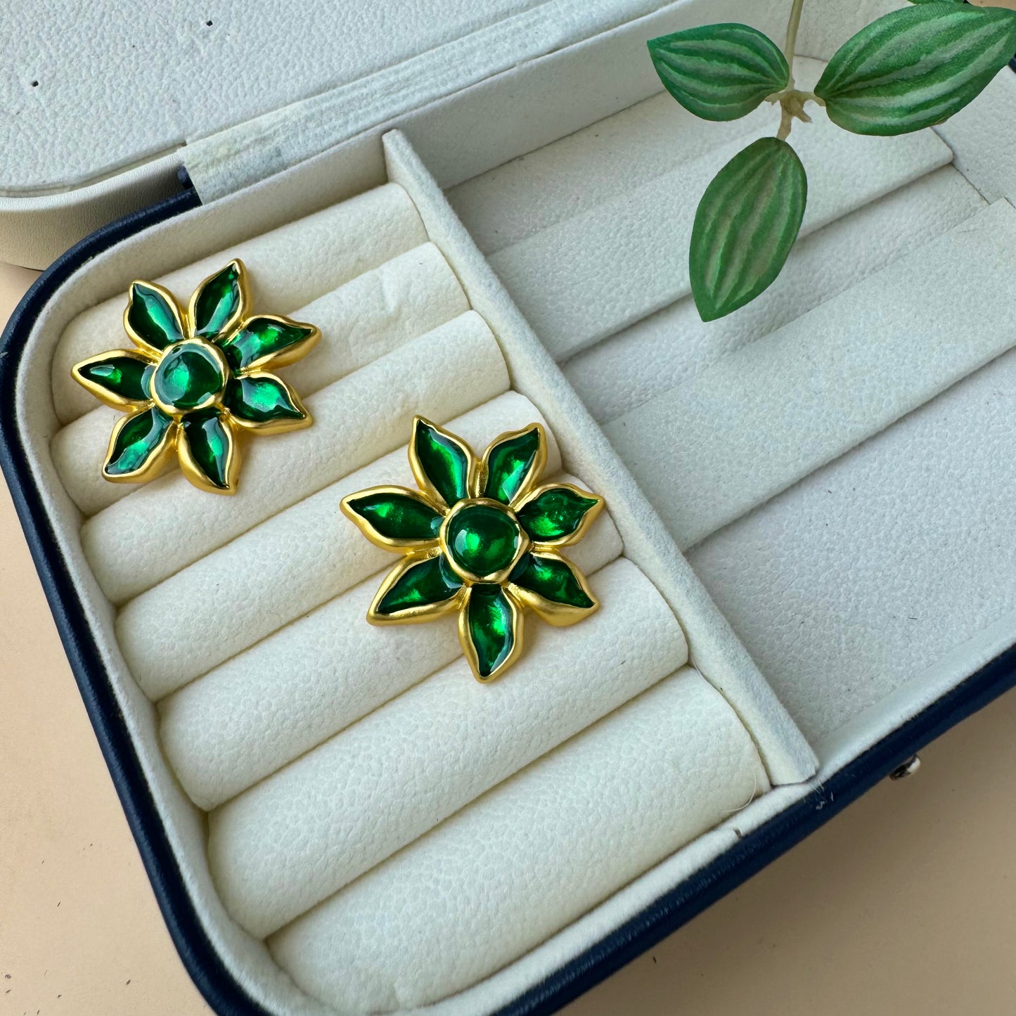 A pair of gold-plated floral earrings with vibrant green enamel petals, displayed on a white cushioned jewelry case. The earrings resemble blooming flowers with a glossy finish, adding an elegant and nature-inspired touch
