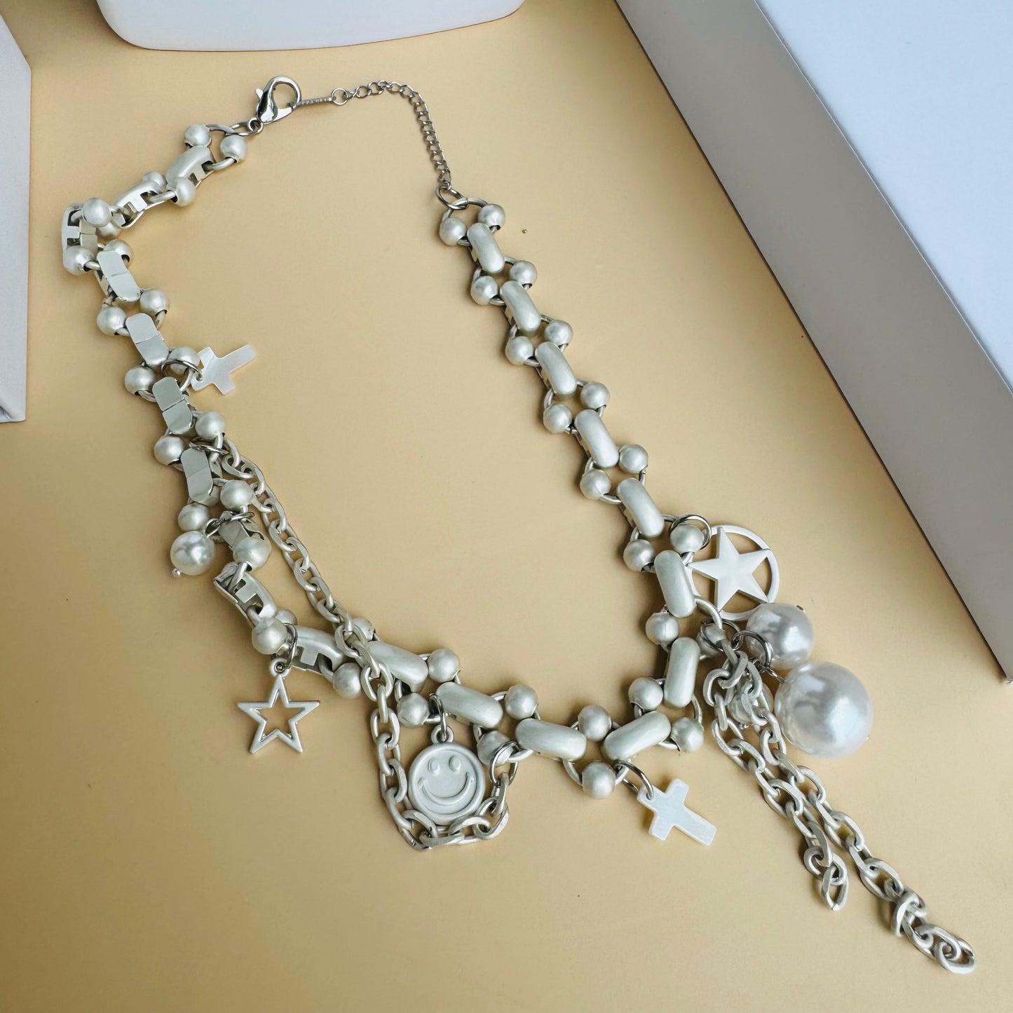 A silver charm necklace displayed on a white bust stand, featuring delicate chains adorned with small charms, including a smiley face, cross, pearl, and abstract shapes. The background includes Lush 24 branded packaging and green plants.