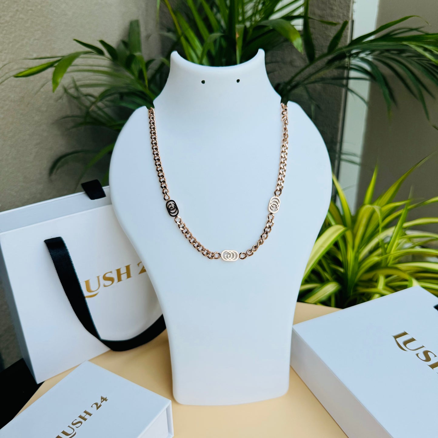 A stylish rose gold chain necklace with delicate interlocking circular accents, displayed on a white jewelry bust with elegant Lush 24 packaging in the background