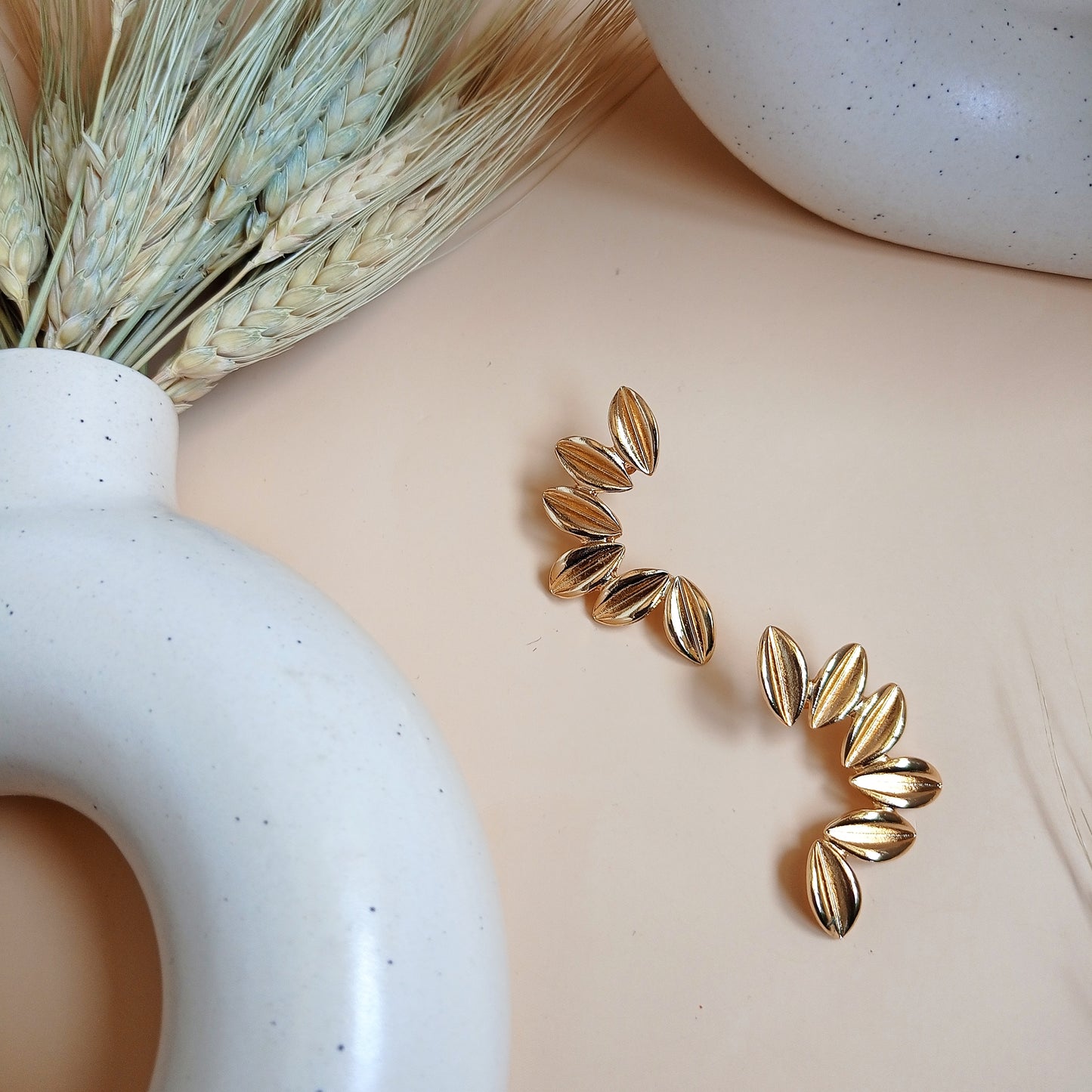 Unique gold statement earrings featuring a curved, leaf-inspired design with a brushed finish, elegantly displayed on a neutral beige background with ceramic and dried floral decor