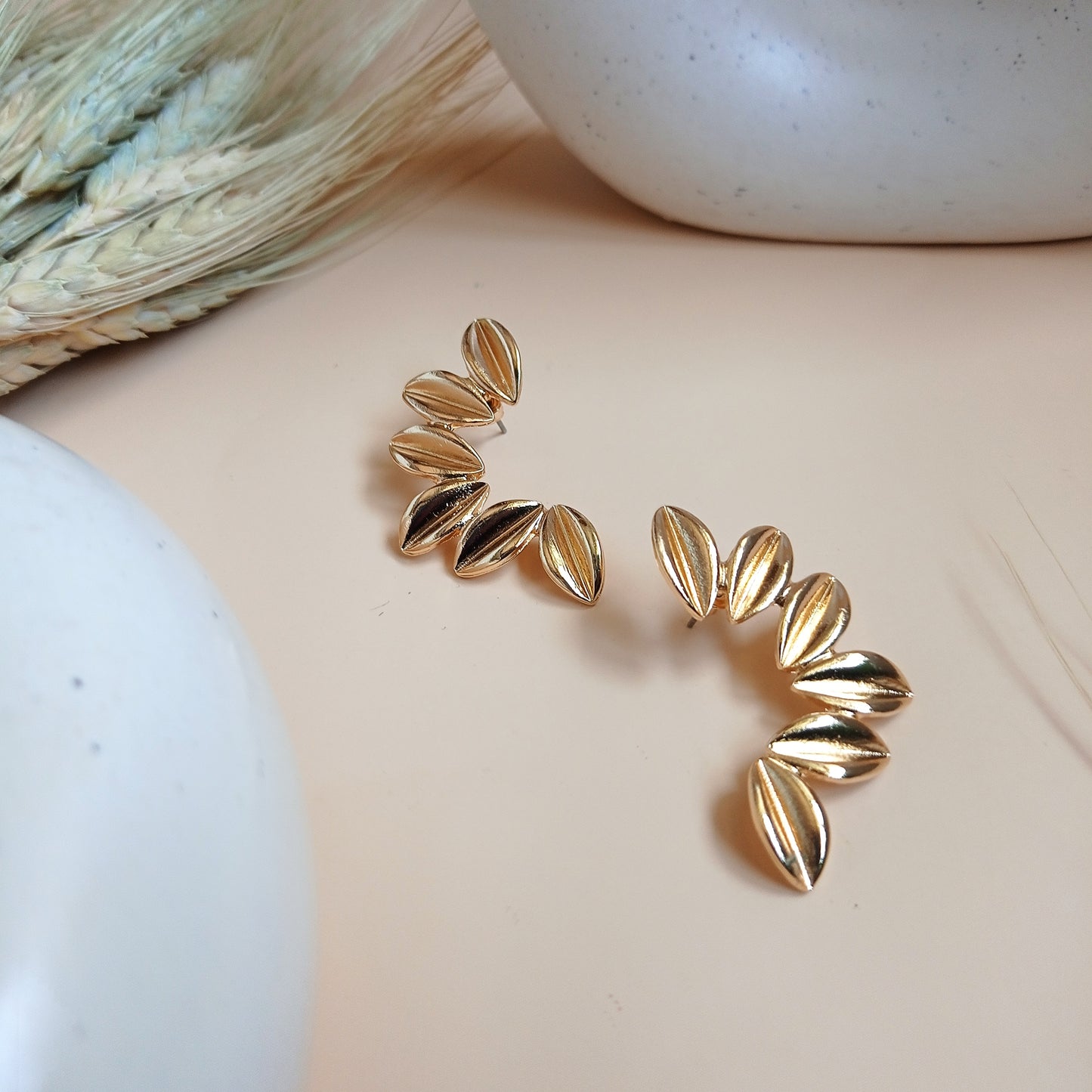 Unique gold statement earrings featuring a curved, leaf-inspired design with a brushed finish, elegantly displayed on a neutral beige background with ceramic and dried floral decor