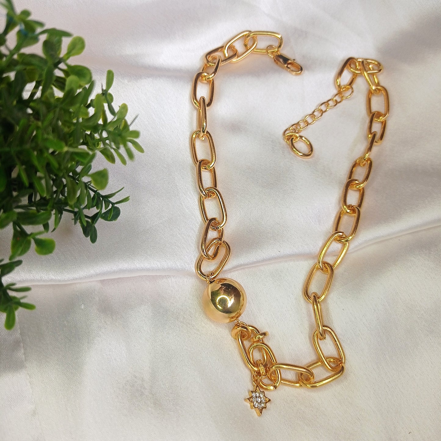 A close-up image of a woman's neck wearing a gold chain necklace with large, elongated links. The necklace features a polished gold sphere and a small dangling star-shaped charm adorned with a sparkling gemstone. The jewelry has a bold and elegant design, standing out against the woman's smooth skin