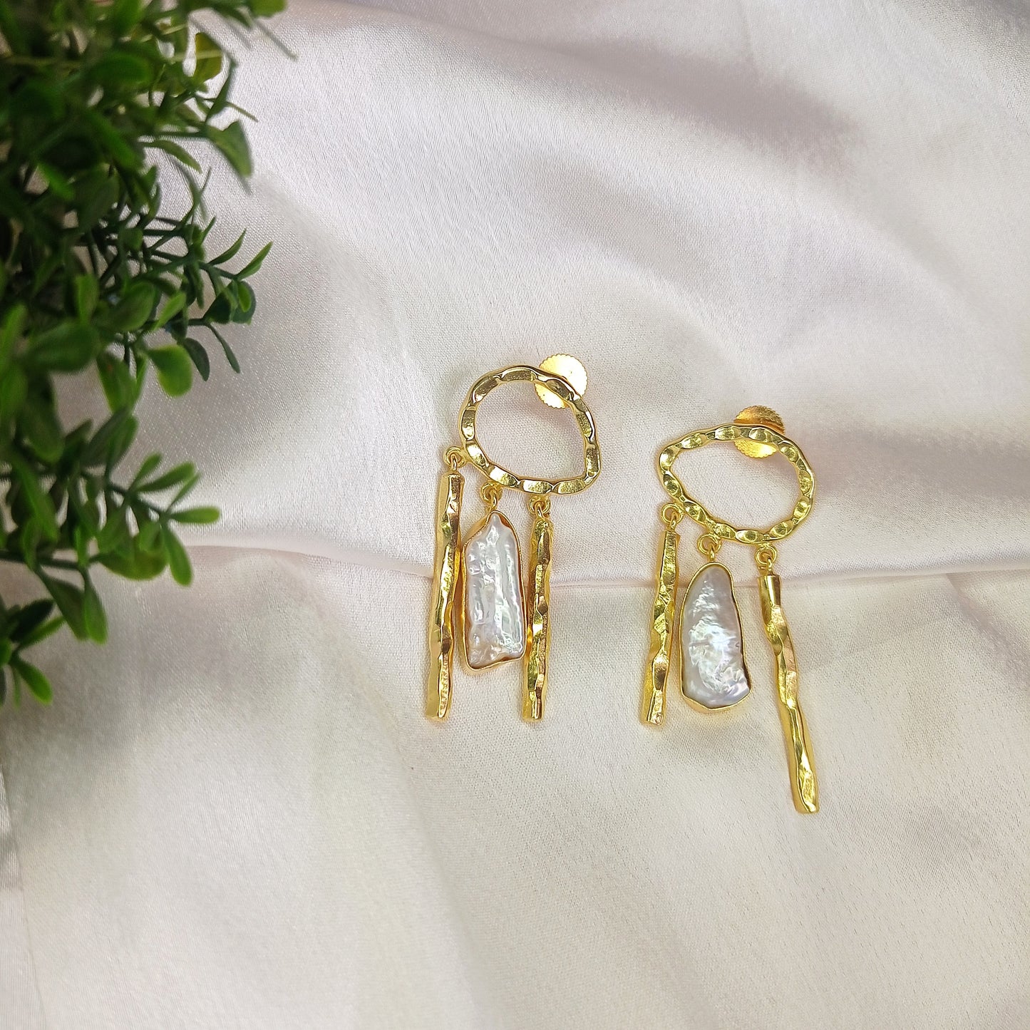 Close-up of a woman wearing artistic gold-tone dangle earrings featuring a hammered open oval stud with cascading organic-shaped bars and a lustrous pearl accent. These bold, sculptural earrings add a touch of modern elegance and sophistication.