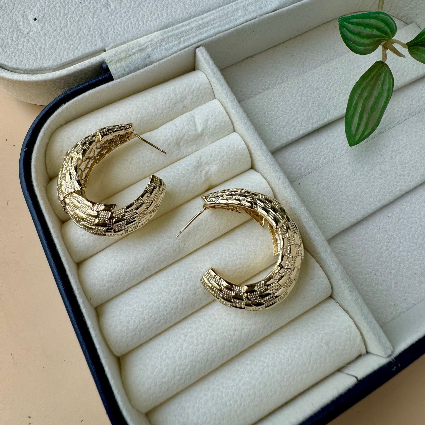 A pair of chunky, textured gold hoop earrings nestled in the soft, white velvet compartments of a dark blue and white jewelry case.