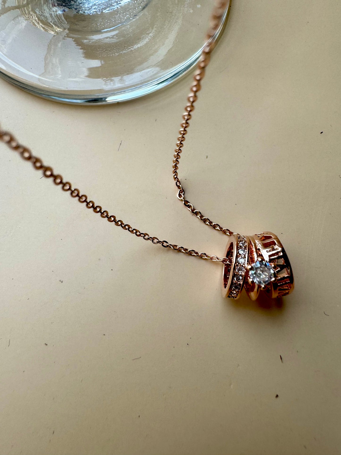 A delicate rose gold chain necklace featuring a pendant composed of stacked rings, one of which is set with clear gemstones resembling diamonds. The pendant has a contemporary, cylindrical design