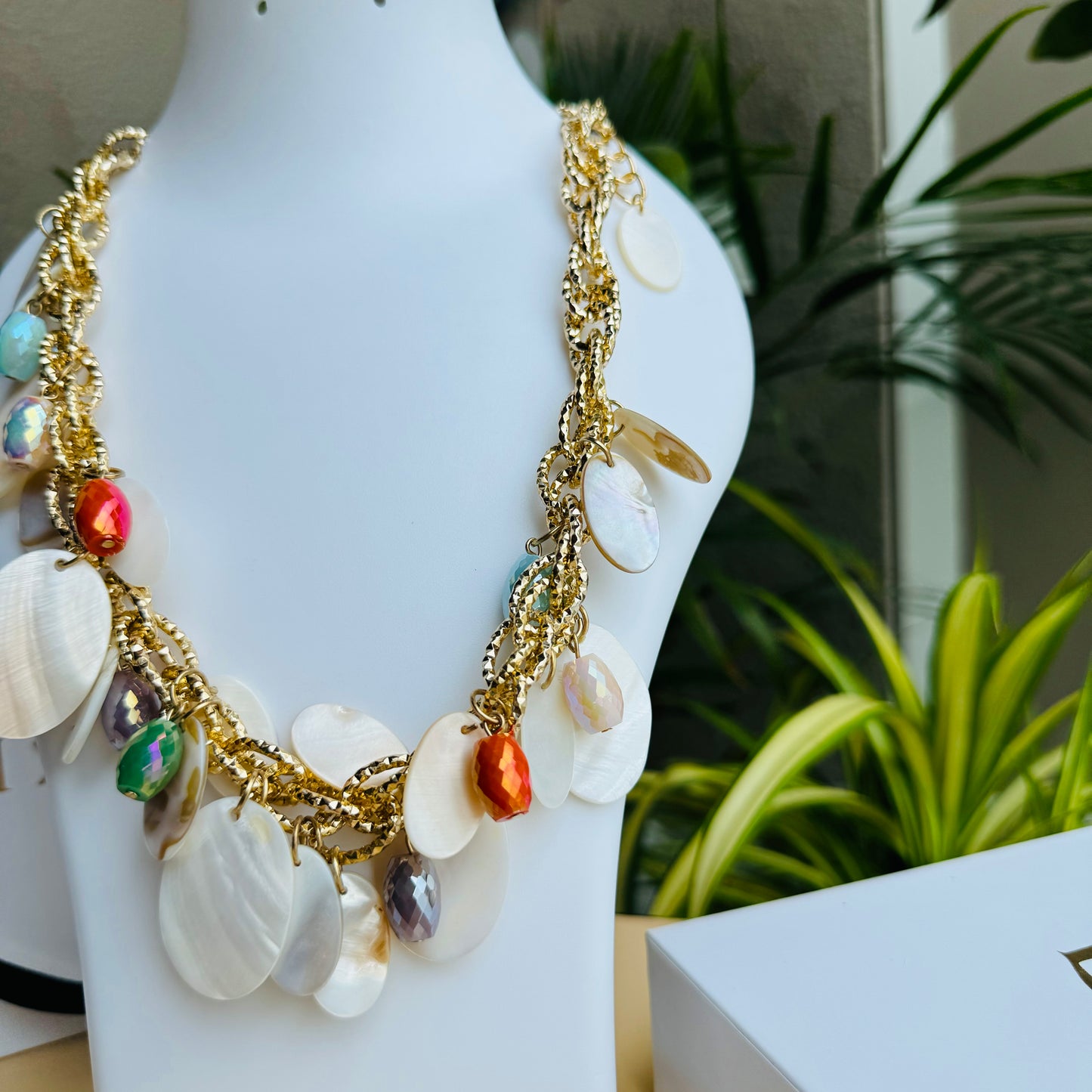 A chunky statement necklace featuring gold-tone chain links adorned with white shell discs and colorful faceted beads. The vibrant mix of orange, blue, and iridescent accents creates a beach-inspired, elegant look. Displayed on a white jewelry bust with a lush green backdrop.