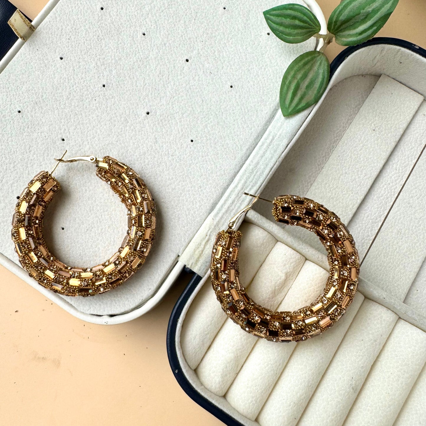 A pair of thick, sparkling gold-toned metal hoop earrings displayed in a light-colored jewelry case with individual compartments. The earrings have a textured surface that catches the light.