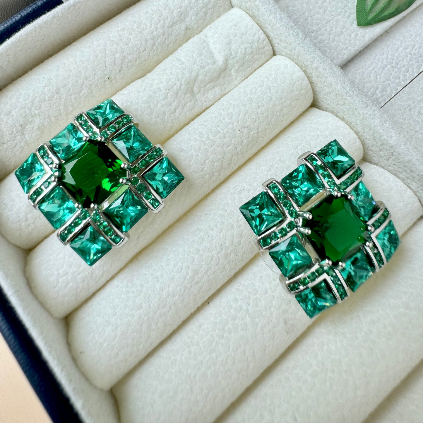 Emerald green square stud earrings with a geometric design, featuring sparkling gemstones set in a silver-tone metal frame, displayed in a cream-colored jewelry box.