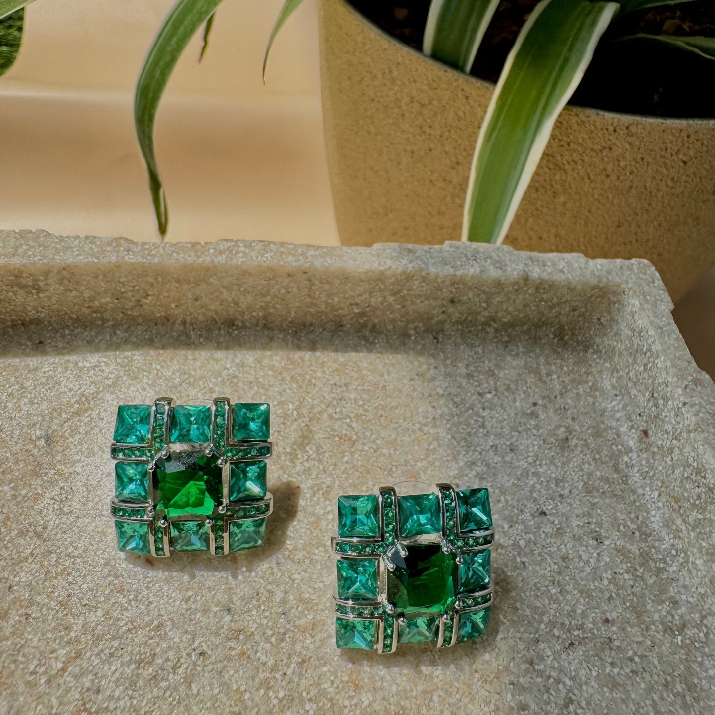 Emerald green square stud earrings with a geometric design, featuring sparkling gemstones set in a silver-tone metal frame, displayed in a cream-colored jewelry box.