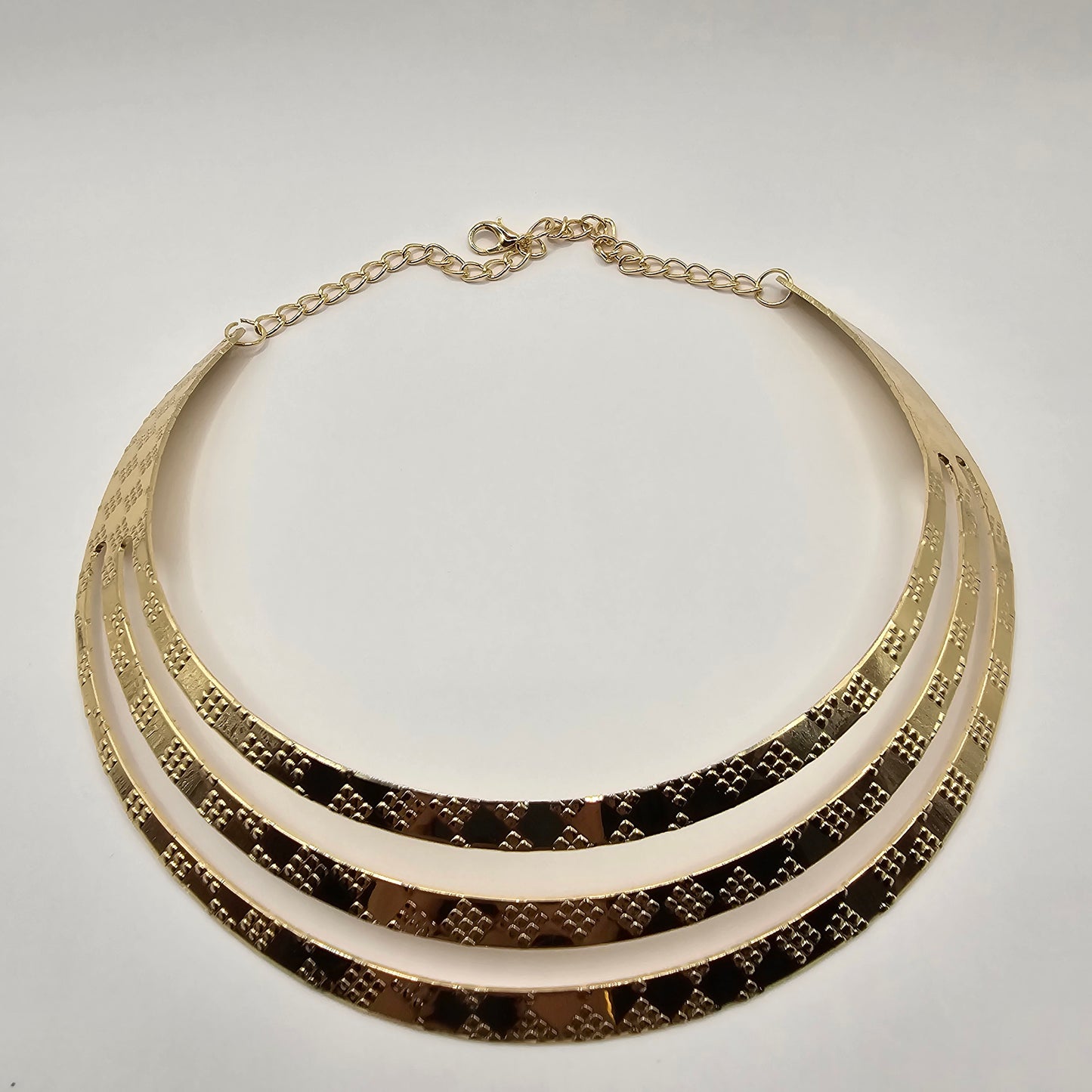 A close-up shot of a large, vintage-inspired gold necklace. The necklace features numerous elongated, slightly curved metallic elements radiating outwards from a central band. It is displayed on a smooth white jewelry bust. In the background, partially visible, are white shopping bags with black handles and the text "LUSH 24" in gold lettering, and a small white jewelry box with the same branding. A blurred view of green tropical foliage is also present.