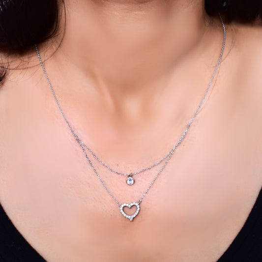 Elegant layered silver necklace featuring a delicate solitaire pendant on the upper chain and a sparkling crystal-studded heart pendant on the lower chain, adding a touch of sophistication and charm
