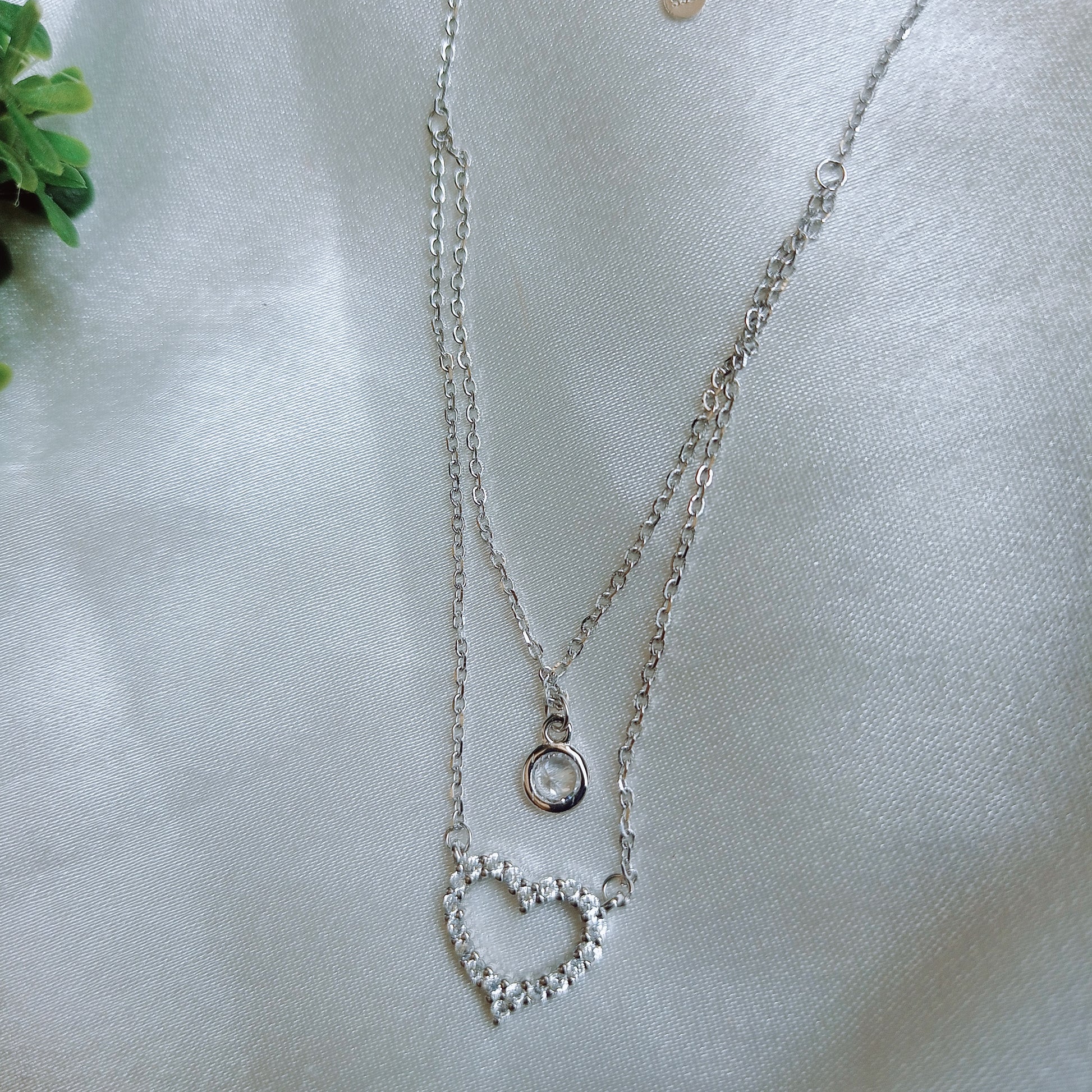 Elegant layered silver necklace featuring a delicate solitaire pendant on the upper chain and a sparkling crystal-studded heart pendant on the lower chain, adding a touch of sophistication and charm