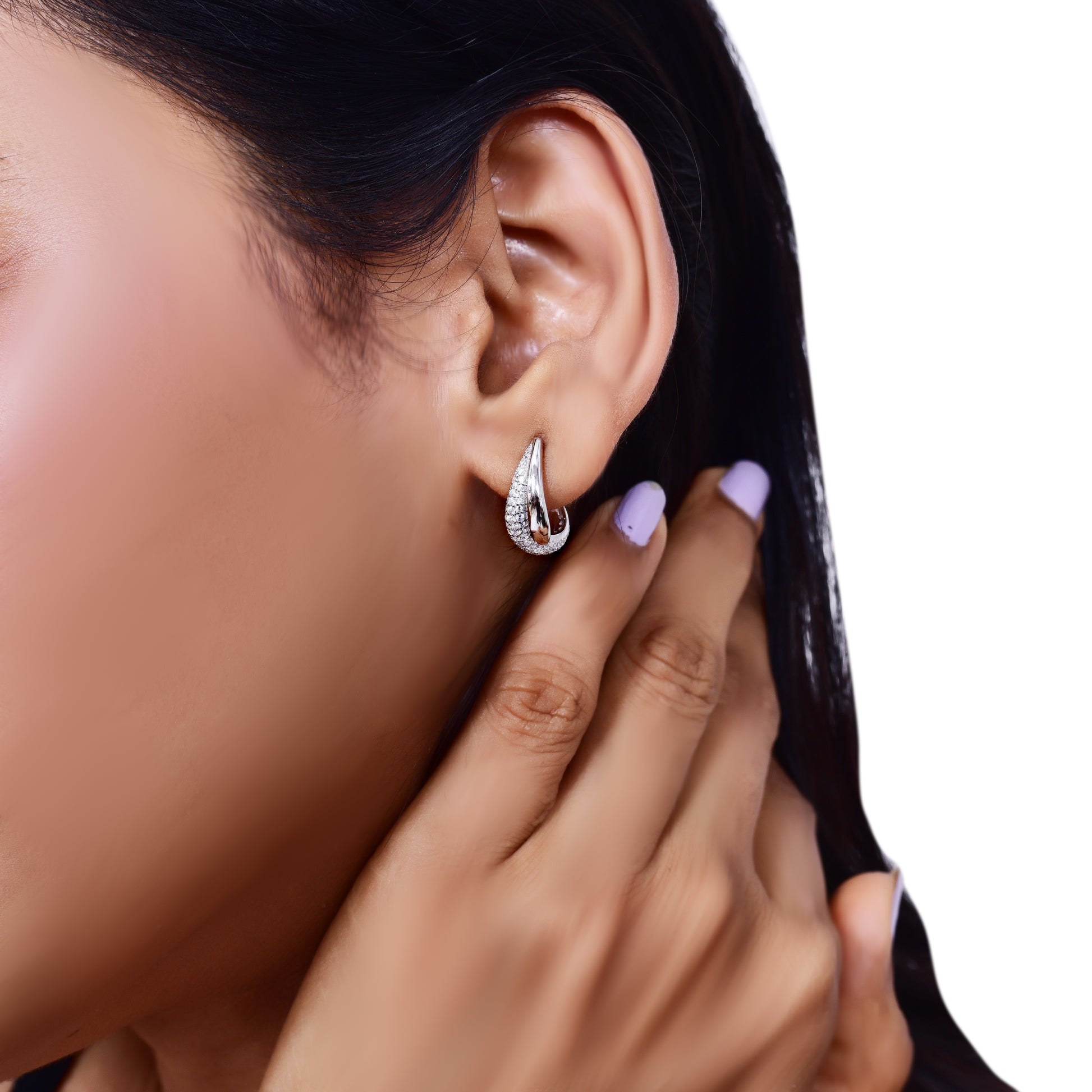The image shows a close-up shot of a pair of silver hoop earrings resting on a soft, white, satin-like fabric. The earrings are small and have a rounded, hoop shape. They appear to be embedded with small, clear stones, possibly diamonds or cubic zirconia, giving them a sparkly appearance.

The fabric is draped softly, creating gentle folds and shadows that add depth to the image. A small portion of green foliage is visible in the bottom left corner, adding a touch of color and natural element to the composi