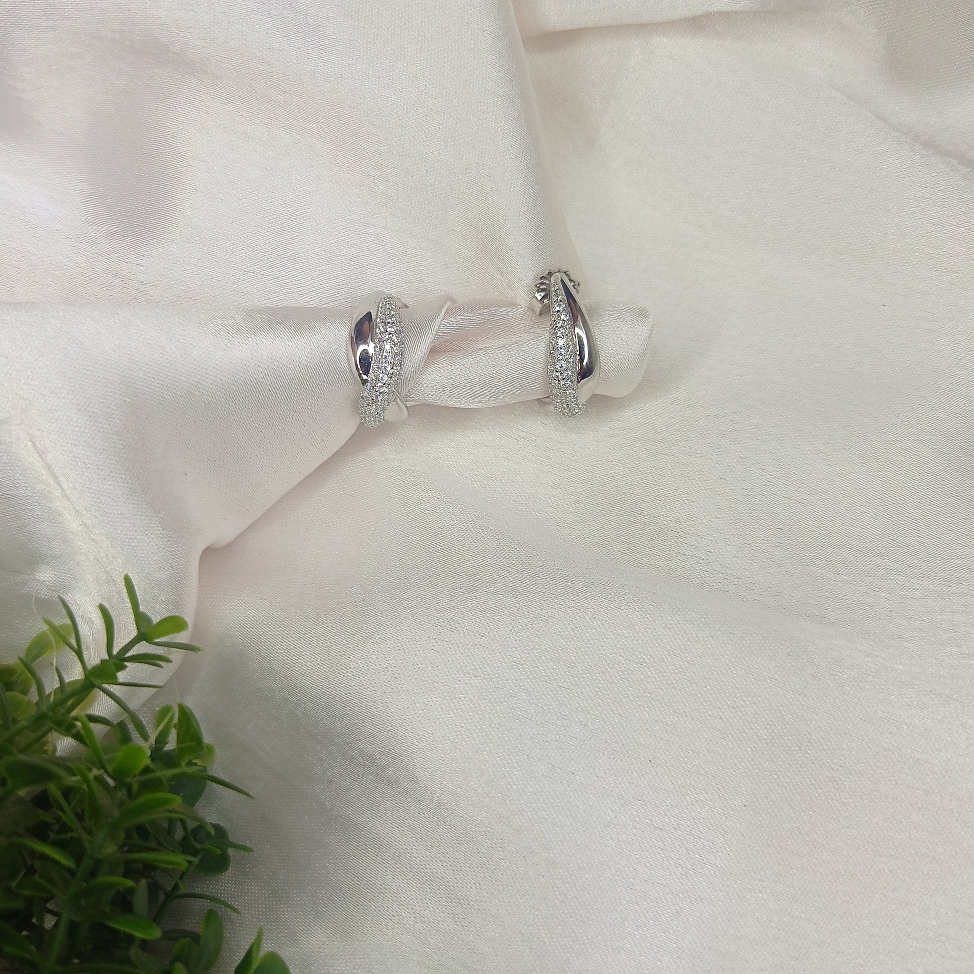 The image shows a close-up shot of a pair of silver hoop earrings resting on a soft, white, satin-like fabric. The earrings are small and have a rounded, hoop shape. They appear to be embedded with small, clear stones, possibly diamonds or cubic zirconia, giving them a sparkly appearance.

The fabric is draped softly, creating gentle folds and shadows that add depth to the image. A small portion of green foliage is visible in the bottom left corner, adding a touch of color and natural element to the composi