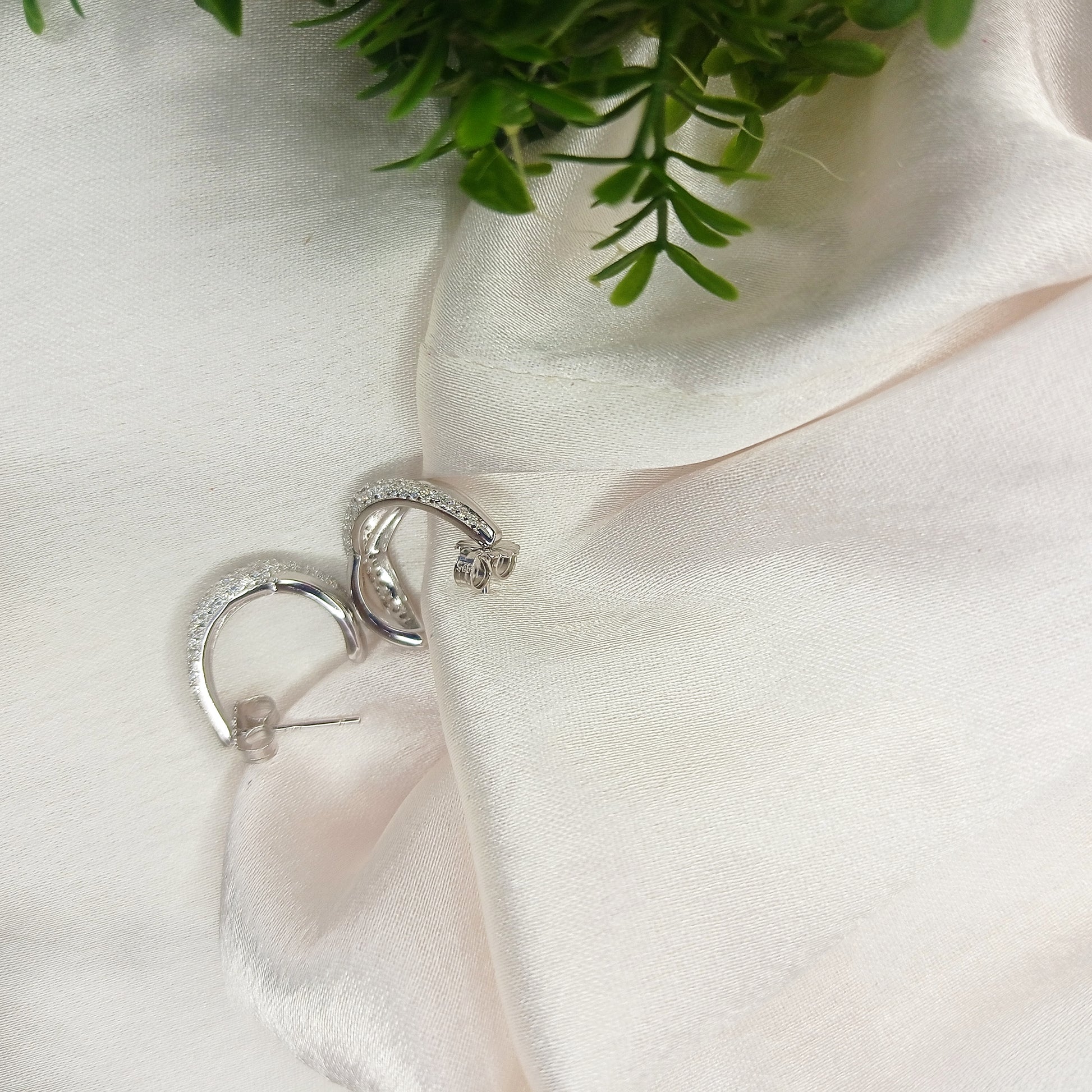 The image shows a close-up shot of a pair of silver hoop earrings resting on a soft, white, satin-like fabric. The earrings are small and have a rounded, hoop shape. They appear to be embedded with small, clear stones, possibly diamonds or cubic zirconia, giving them a sparkly appearance.

The fabric is draped softly, creating gentle folds and shadows that add depth to the image. A small portion of green foliage is visible in the bottom left corner, adding a touch of color and natural element to the composi