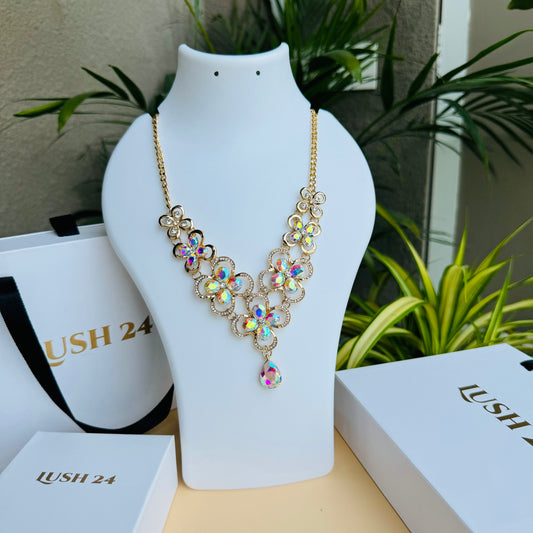 Gold statement necklace with iridescent floral-shaped gemstones and a teardrop pendant, displayed on a white mannequin bust. The intricate design sparkles with multicolored reflections, creating a luxurious and elegant look