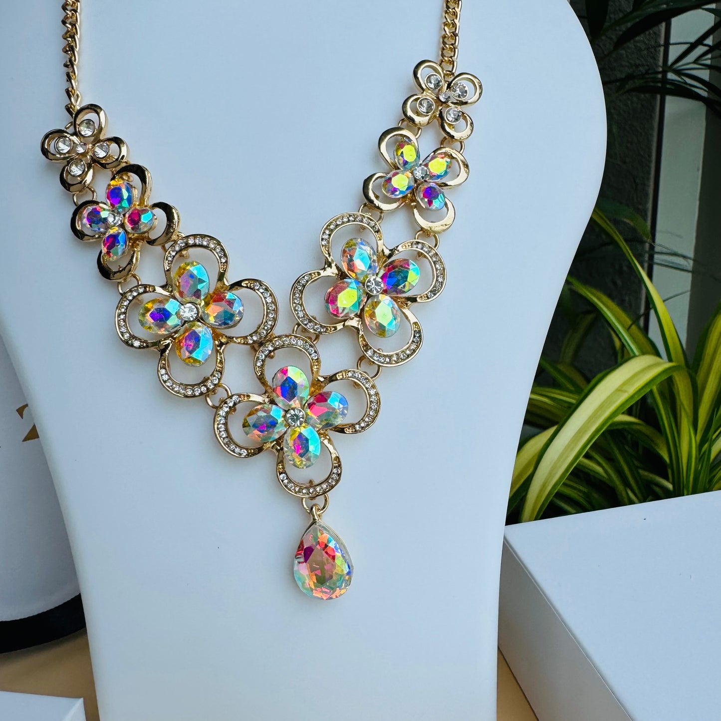 Gold statement necklace with iridescent floral-shaped gemstones and a teardrop pendant, displayed on a white mannequin bust. The intricate design sparkles with multicolored reflections, creating a luxurious and elegant look