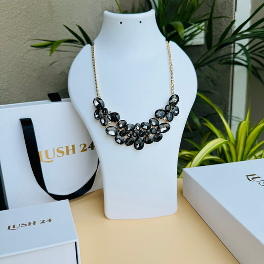Elegant statement necklace with black and smoky crystal stones on a gold chain, displayed on a white mannequin bust. Branded Lush 24 packaging in the background adds a premium touch.