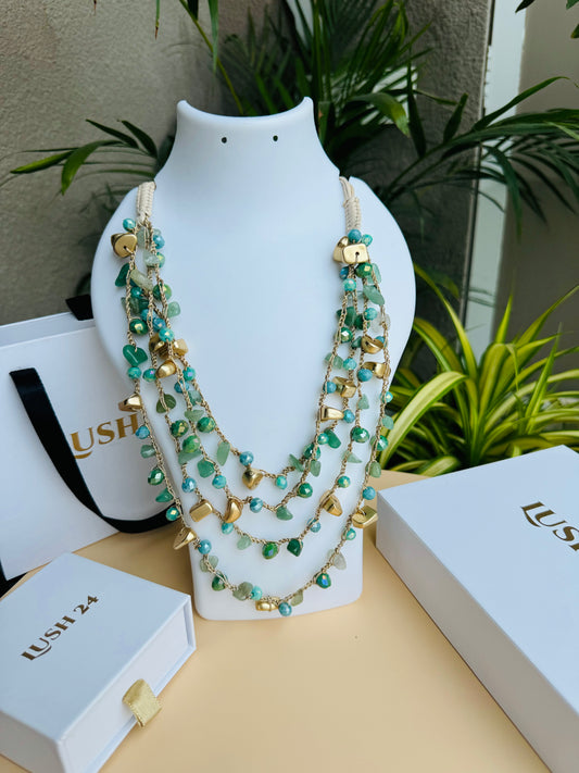 A multi-layered necklace with blue-green glass beads and gold accents displayed on a white bust stand. The necklace features delicate strands of beads interwoven with small golden charms. The background includes lush green plants and branded Lush 24 packaging boxes