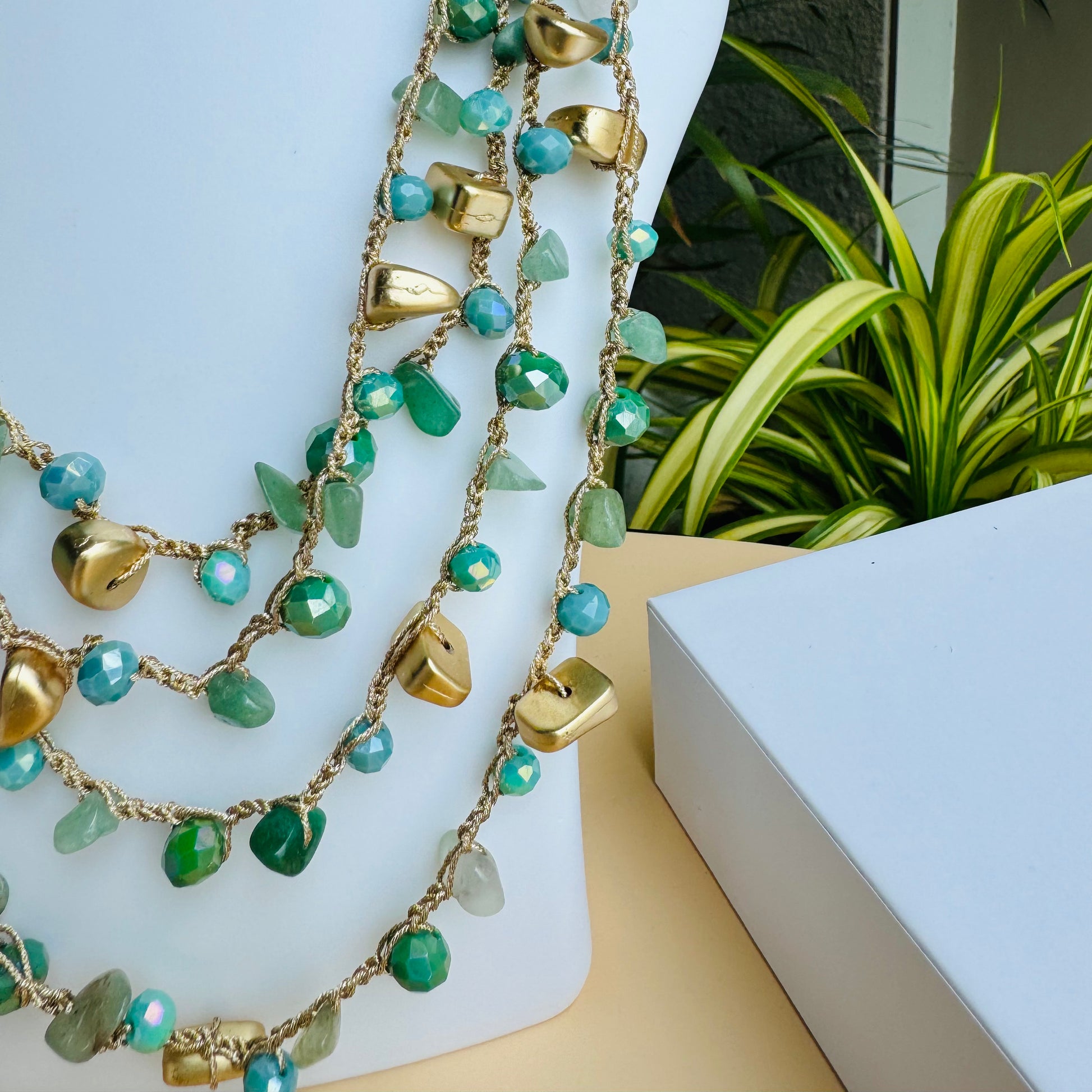 A multi-layered necklace with blue-green glass beads and gold accents displayed on a white bust stand. The necklace features delicate strands of beads interwoven with small golden charms. The background includes lush green plants and branded Lush 24 packaging boxes