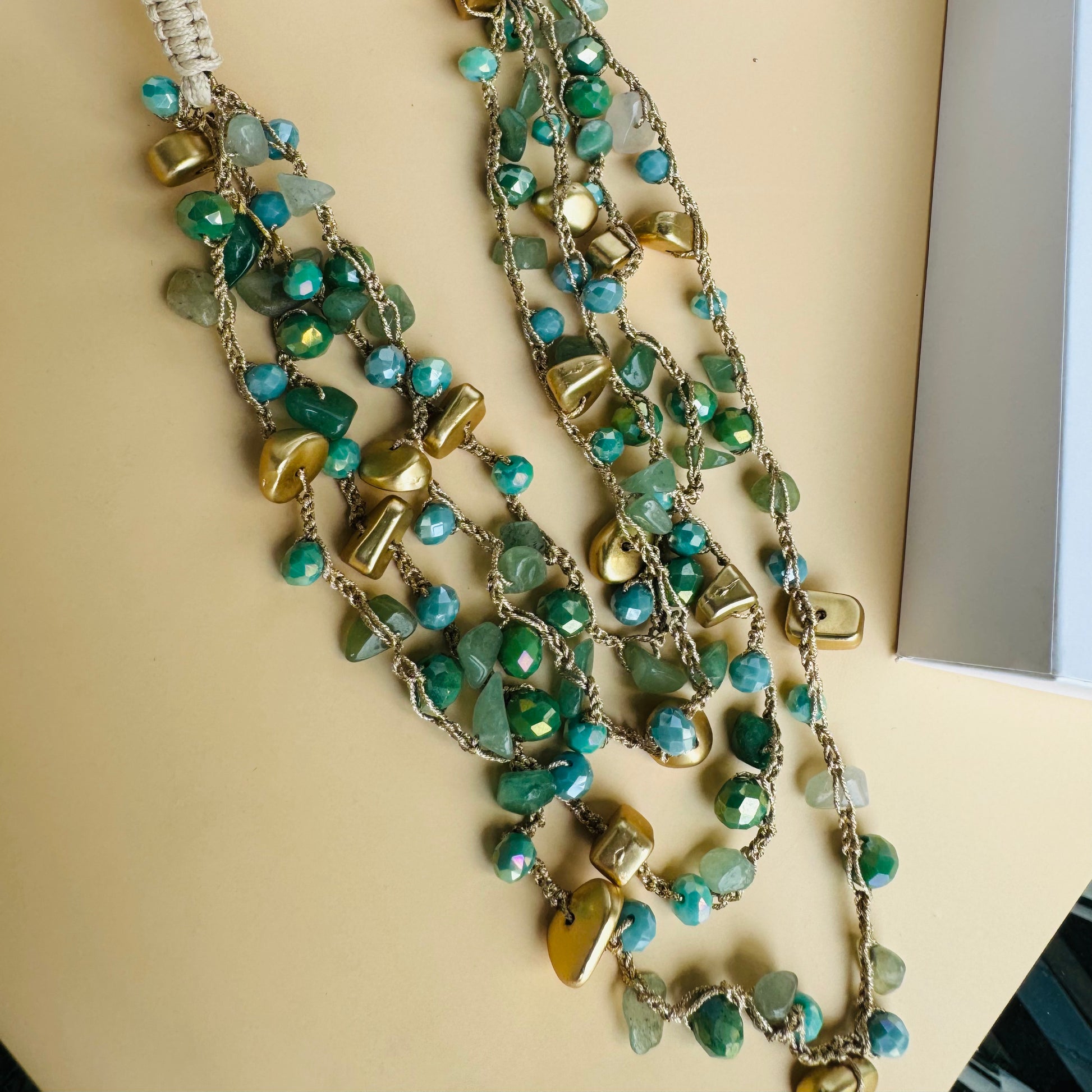 A multi-layered necklace with blue-green glass beads and gold accents displayed on a white bust stand. The necklace features delicate strands of beads interwoven with small golden charms. The background includes lush green plants and branded Lush 24 packaging boxes