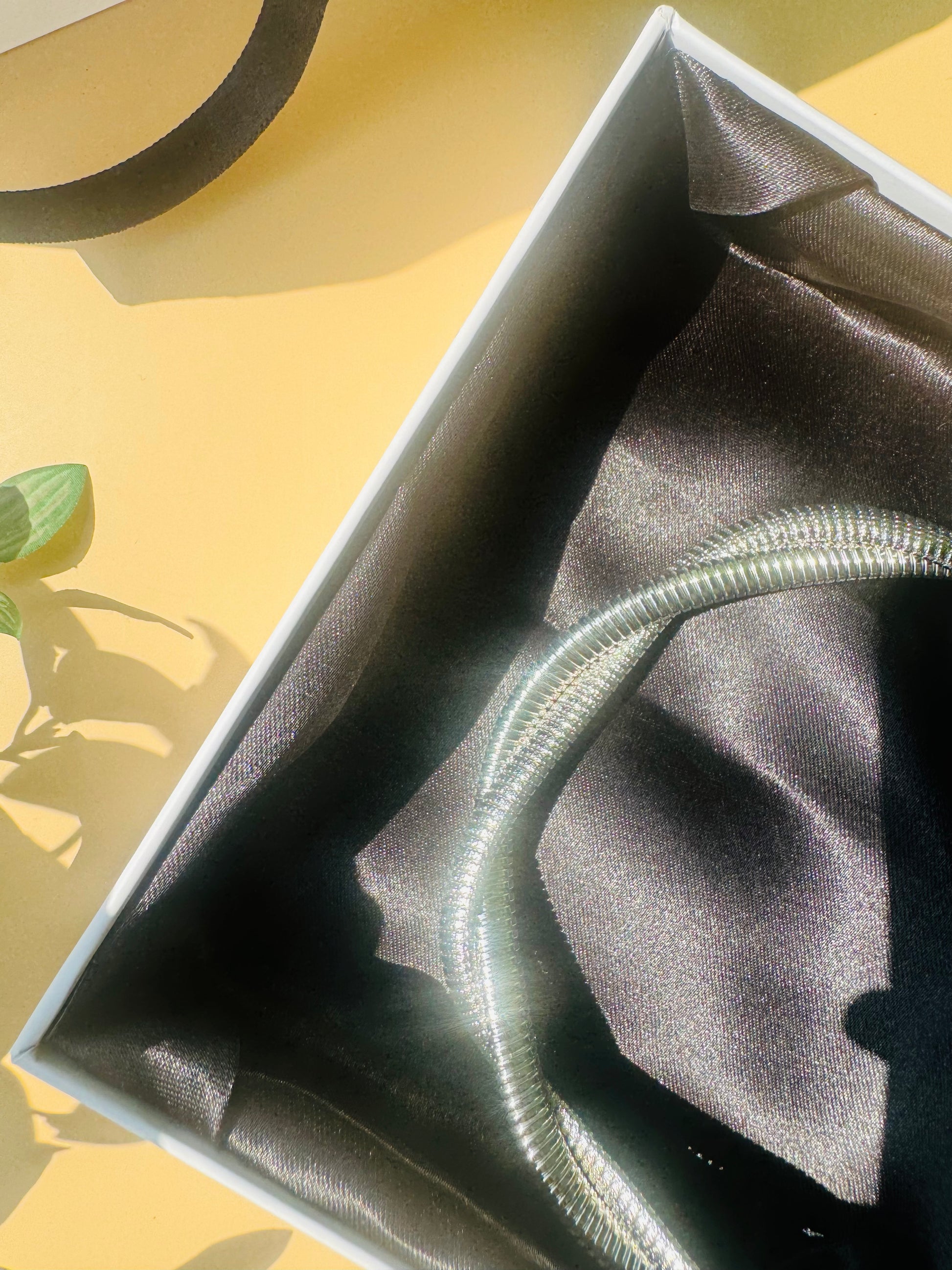 A sleek and modern silver-toned necklace with a coiled, tubular design, elegantly displayed on a white bust stand, with luxury packaging in the background
