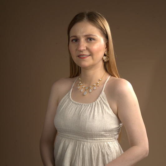 Smiling woman wearing a cream-colored sleeveless dress, accessorized with a gold and white gemstone statement necklace and matching dangle earrings against a warm brown background