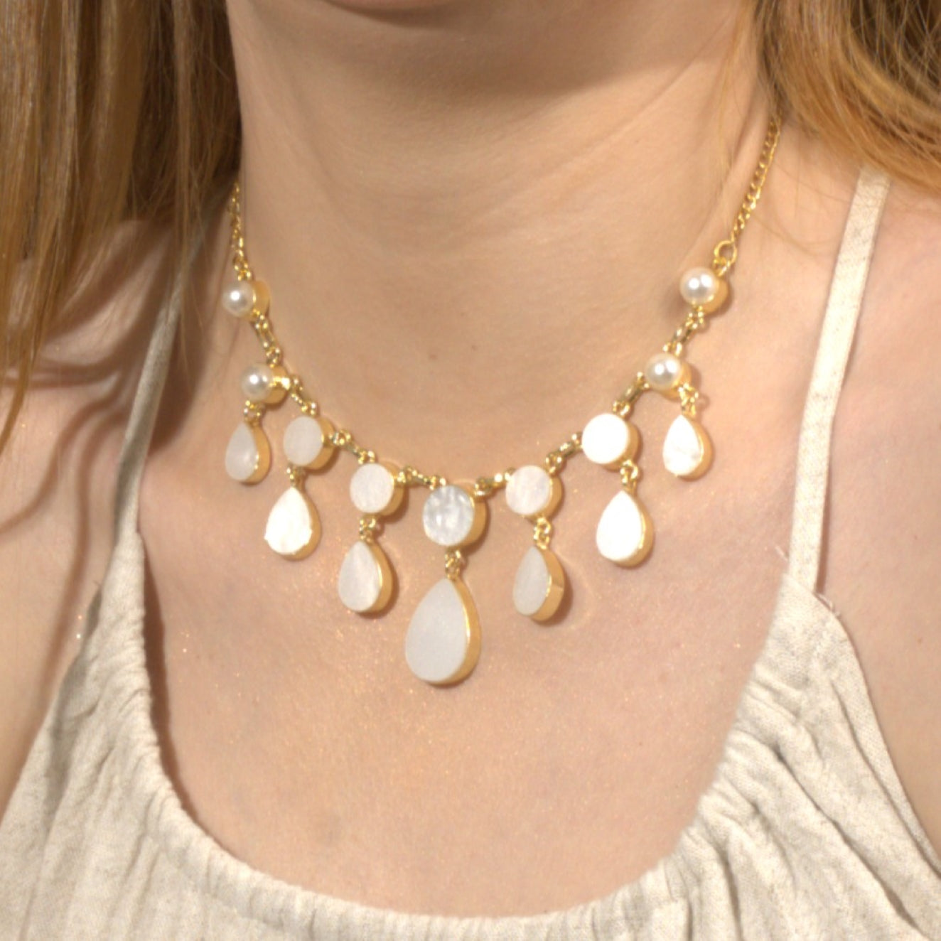Smiling woman wearing a cream-colored sleeveless dress, accessorized with a gold and white gemstone statement necklace and matching dangle earrings against a warm brown background