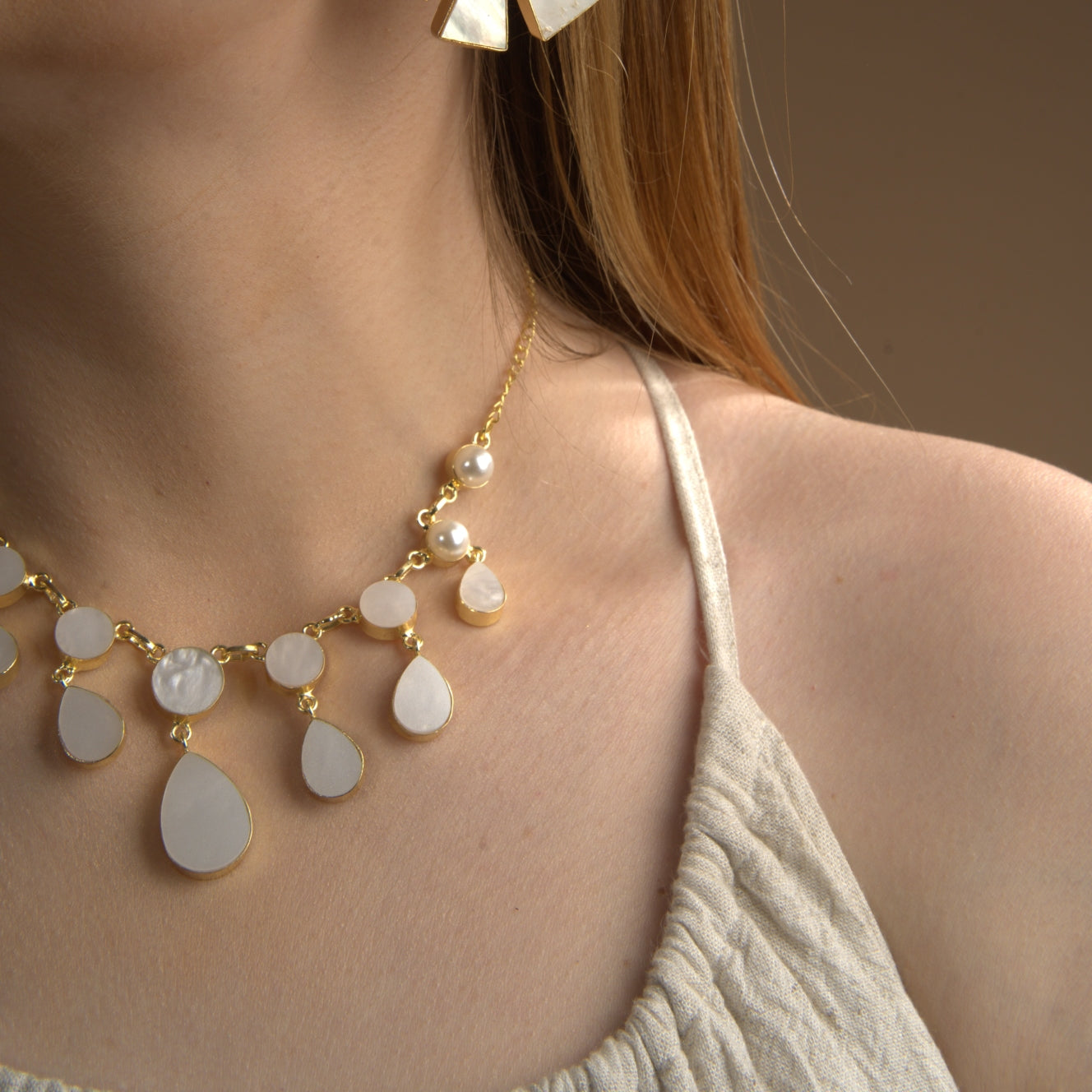 Smiling woman wearing a cream-colored sleeveless dress, accessorized with a gold and white gemstone statement necklace and matching dangle earrings against a warm brown background