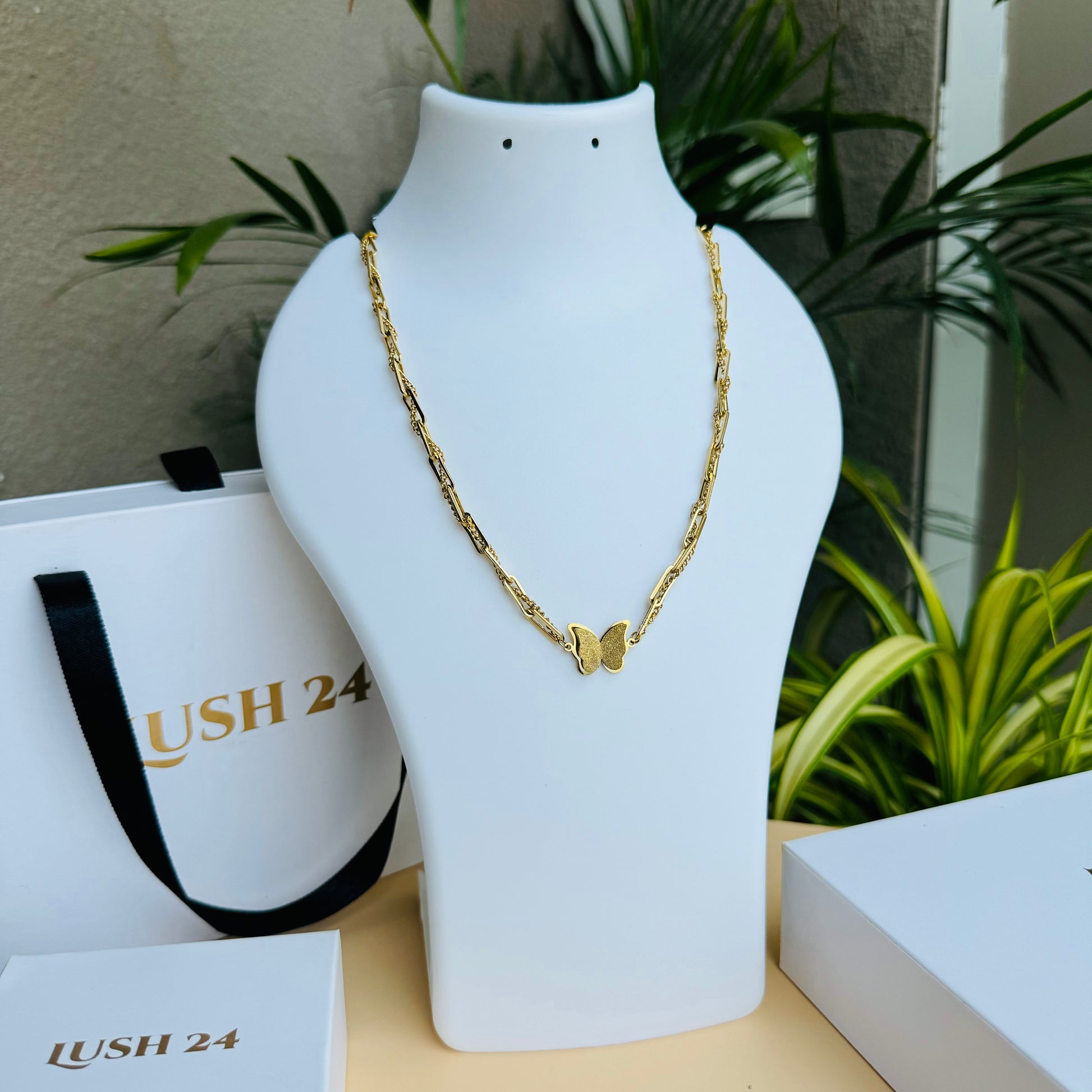 A delicate gold chain necklace featuring a butterfly pendant at its center. The chain has an intricate design, adding elegance to the minimalistic yet stylish piece. Displayed on a white mannequin bust with Lush 24 packaging in the background, creating a luxurious presentation.