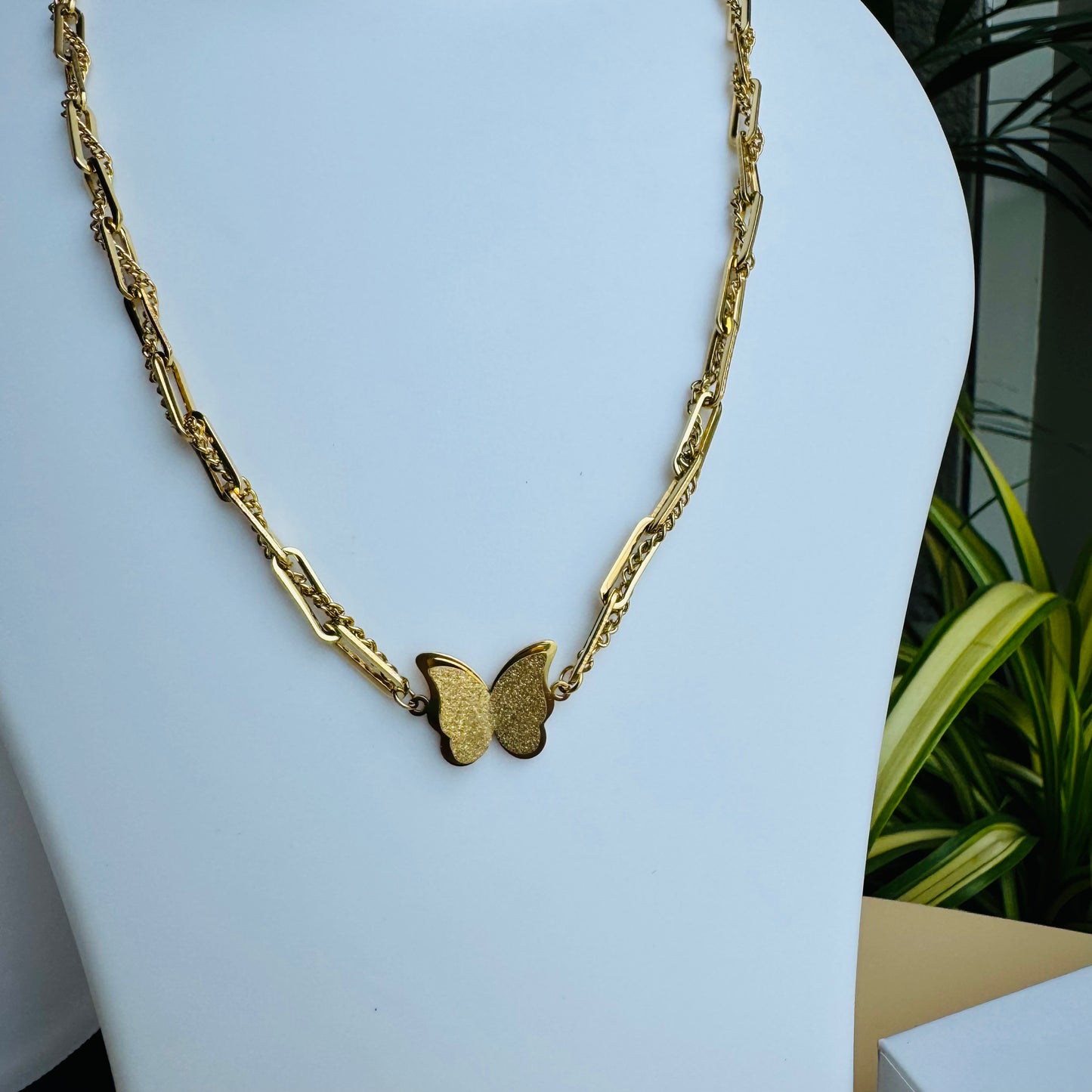 A delicate gold chain necklace featuring a butterfly pendant at its center. The chain has an intricate design, adding elegance to the minimalistic yet stylish piece. Displayed on a white mannequin bust with Lush 24 packaging in the background, creating a luxurious presentation.