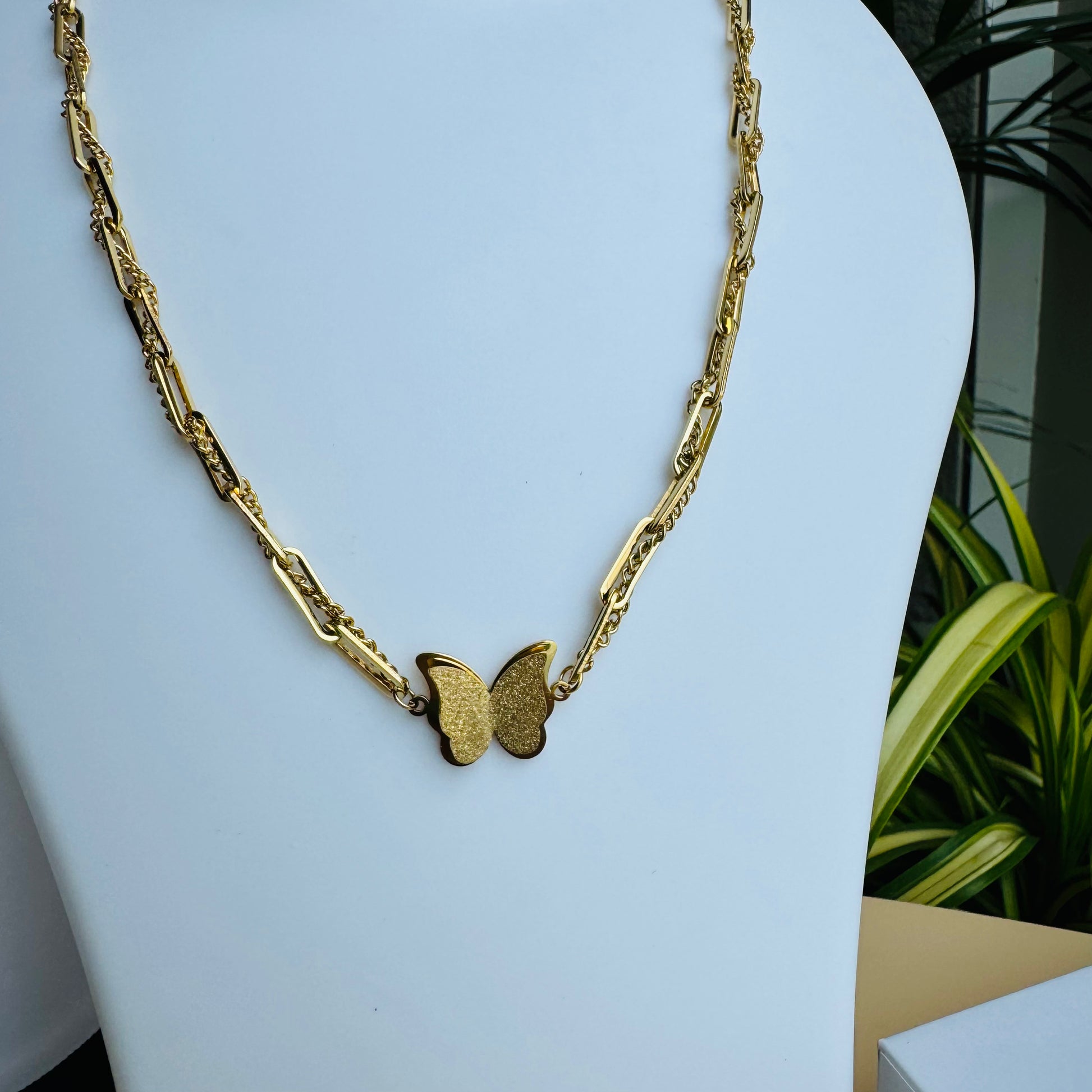 A delicate gold chain necklace featuring a butterfly pendant at its center. The chain has an intricate design, adding elegance to the minimalistic yet stylish piece. Displayed on a white mannequin bust with Lush 24 packaging in the background, creating a luxurious presentation.