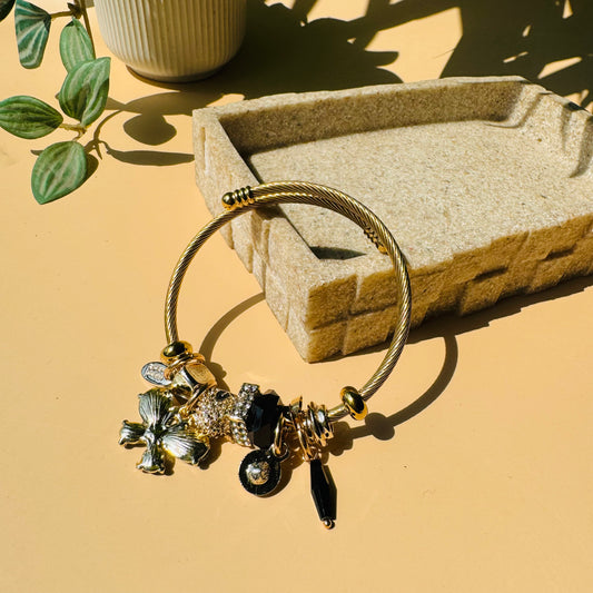 A Pandora-style charm bracelet with a gold-toned wire bangle and a collection of charms in mixed metals, including a silver-toned flower, a pave crystal bead, and black and gold accents. Likely available in Mumbai, Maharashtra, India.