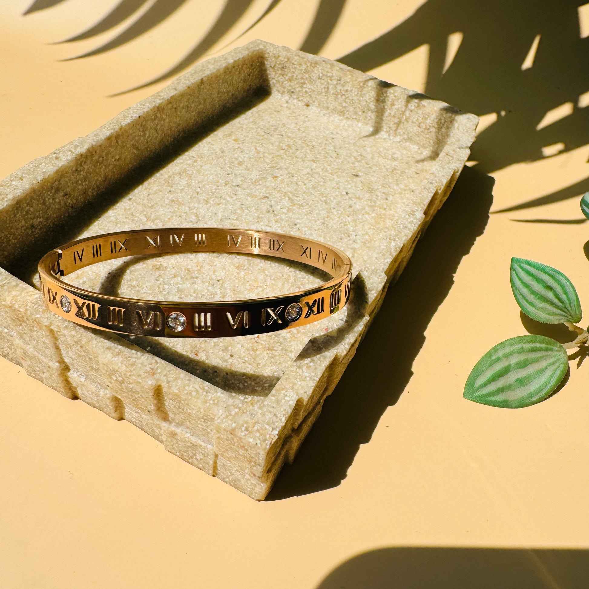 Authentic gold roman numerals Bracelet. The bracelet is showcased on a textured stone-like block with soft lighting and a glimpse of plant life.
