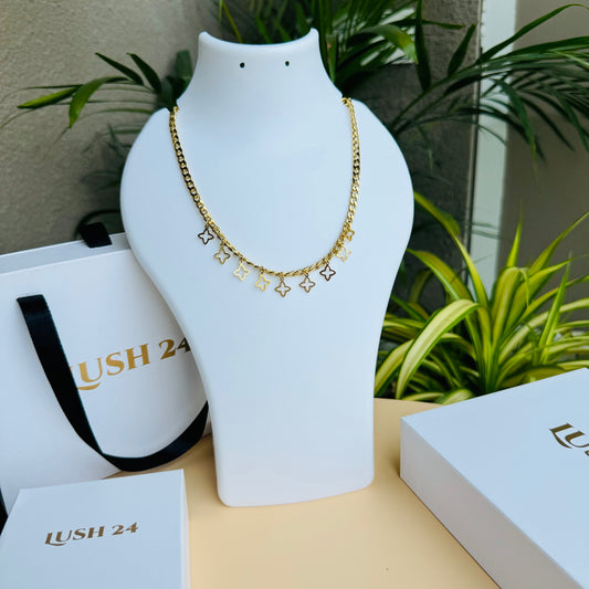 This is a gold chain necklace adorned with small dangling star-shaped charms. The elegant design adds a delicate yet trendy touch to the piece. Displayed on a white mannequin bust, the necklace is complemented by Lush 24 packaging in the background, enhancing its luxury appeal.