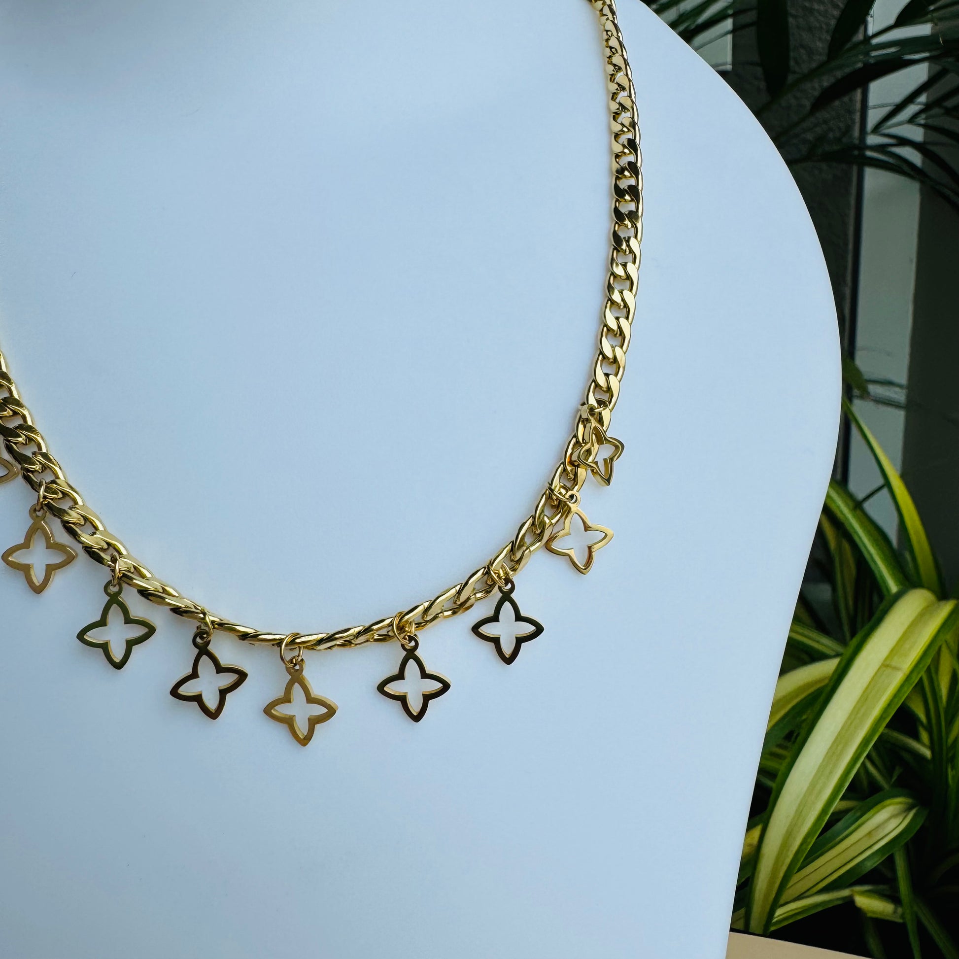 This is a gold chain necklace adorned with small dangling star-shaped charms. The elegant design adds a delicate yet trendy touch to the piece. Displayed on a white mannequin bust, the necklace is complemented by Lush 24 packaging in the background, enhancing its luxury appeal.