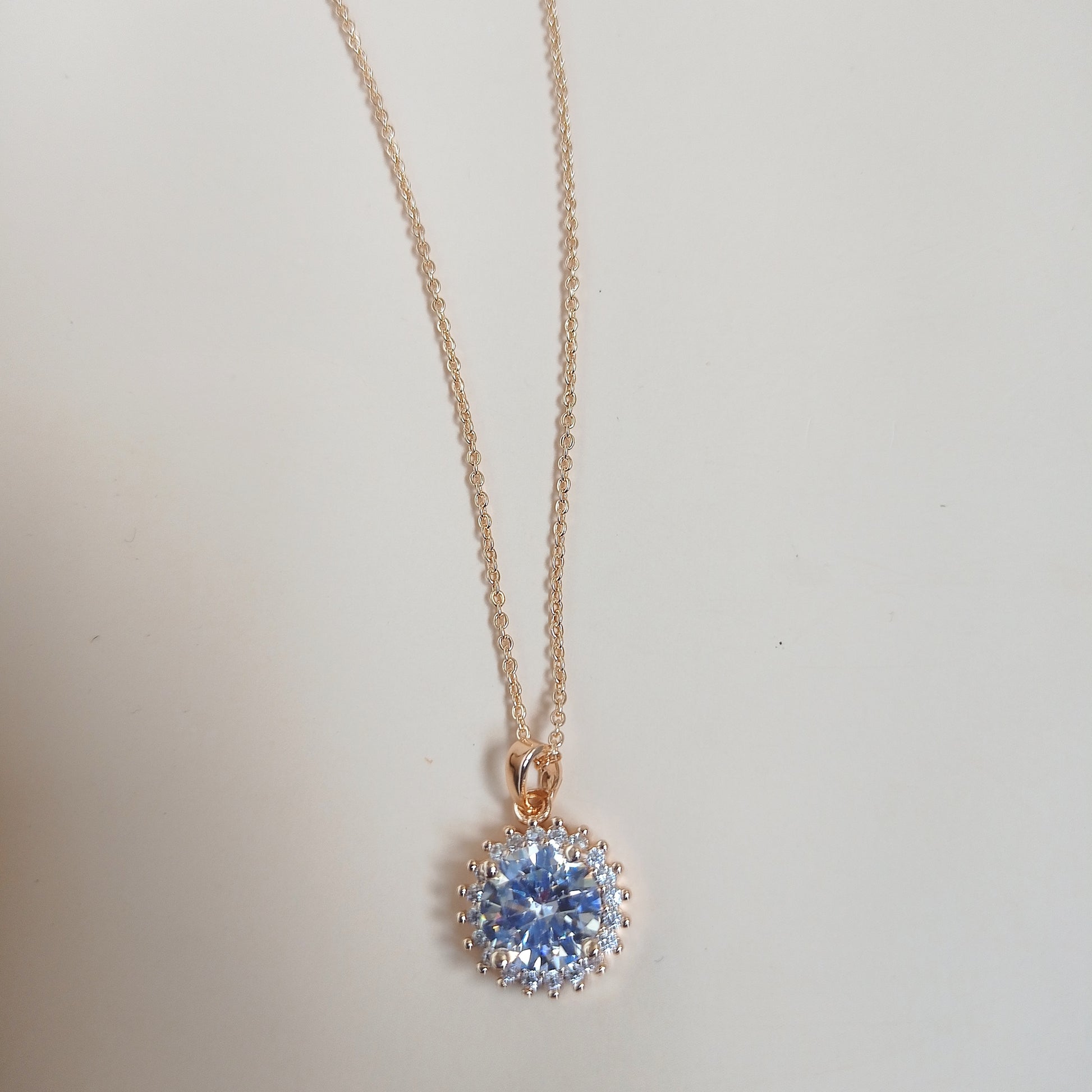 Elegant gold chain necklace featuring a dazzling round-cut blue gemstone pendant, surrounded by a halo of smaller stones, set against a neutral background